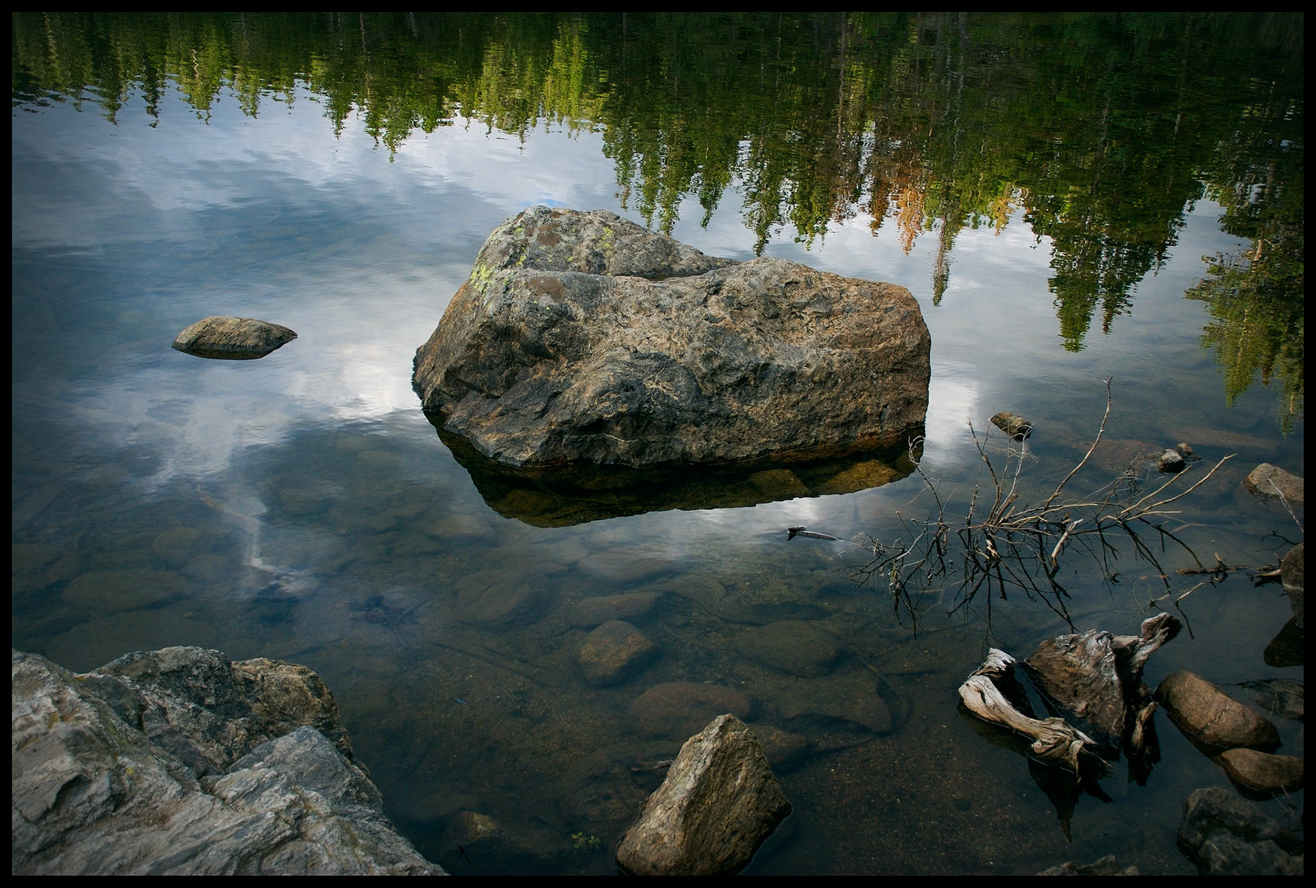 Boulder in Bear Lake, Rocky Mountain National Park, Colorado USA with pine forest and cloudy sky reflected in water.