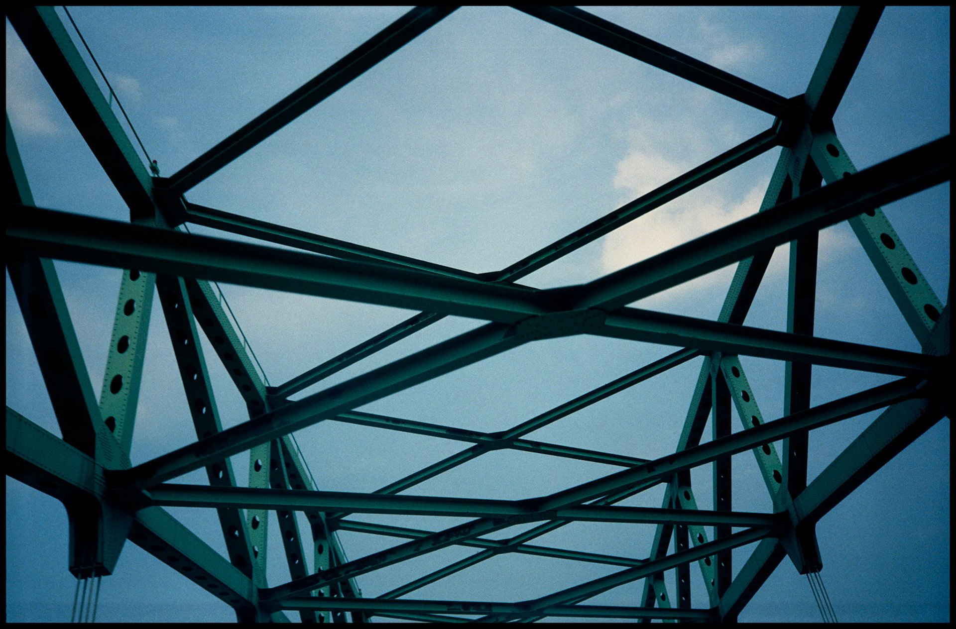 An upward view of the angular patterns created by green iron bridge trusses against a pale blue sky. Kansas City, Missouri, 1991