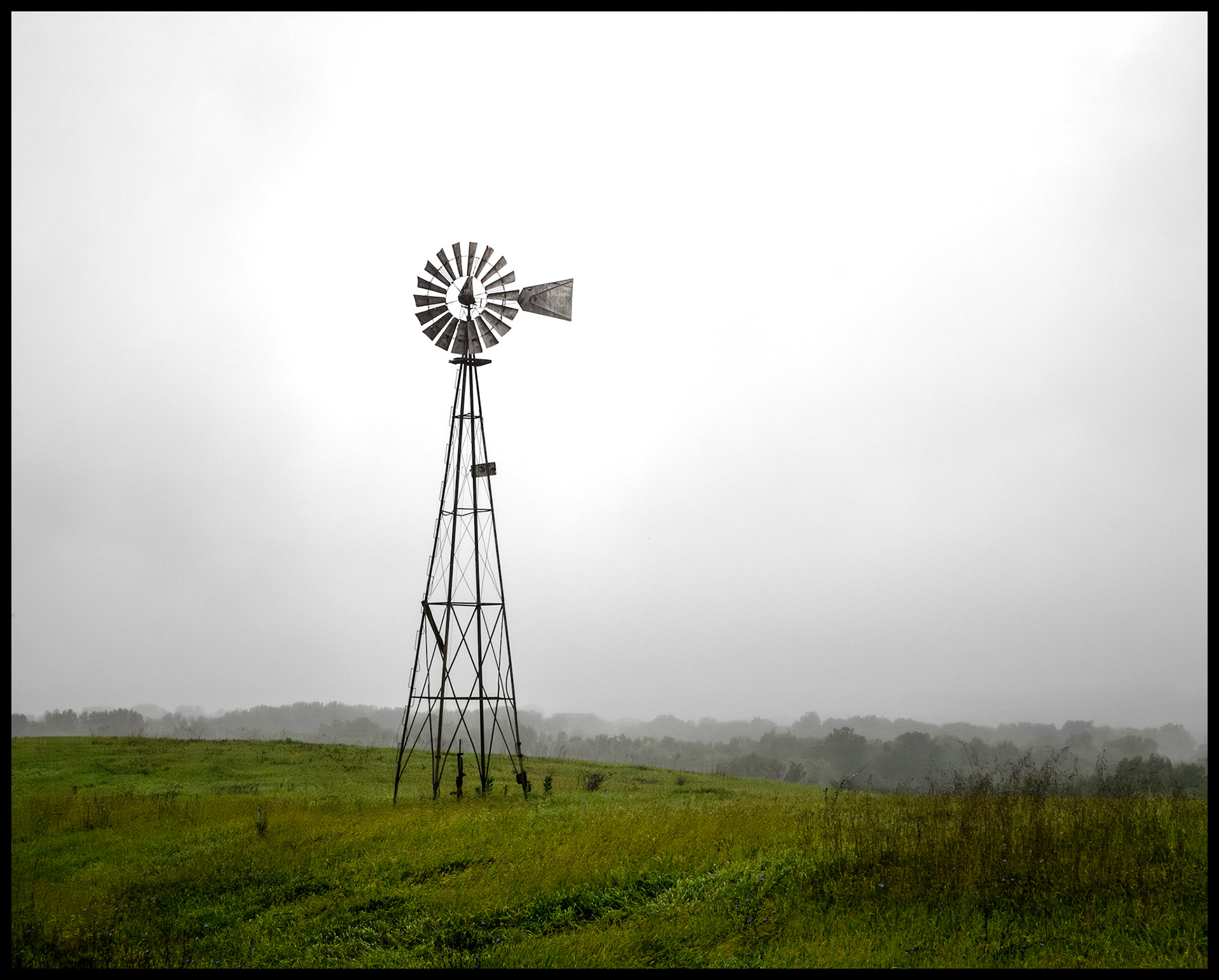 A solitary windmill at the edge of a green field silhoutted against the overcast sky on a rainy and foggy September morning. Located on Indian Hill near South Gifford, Missouri, 2023.