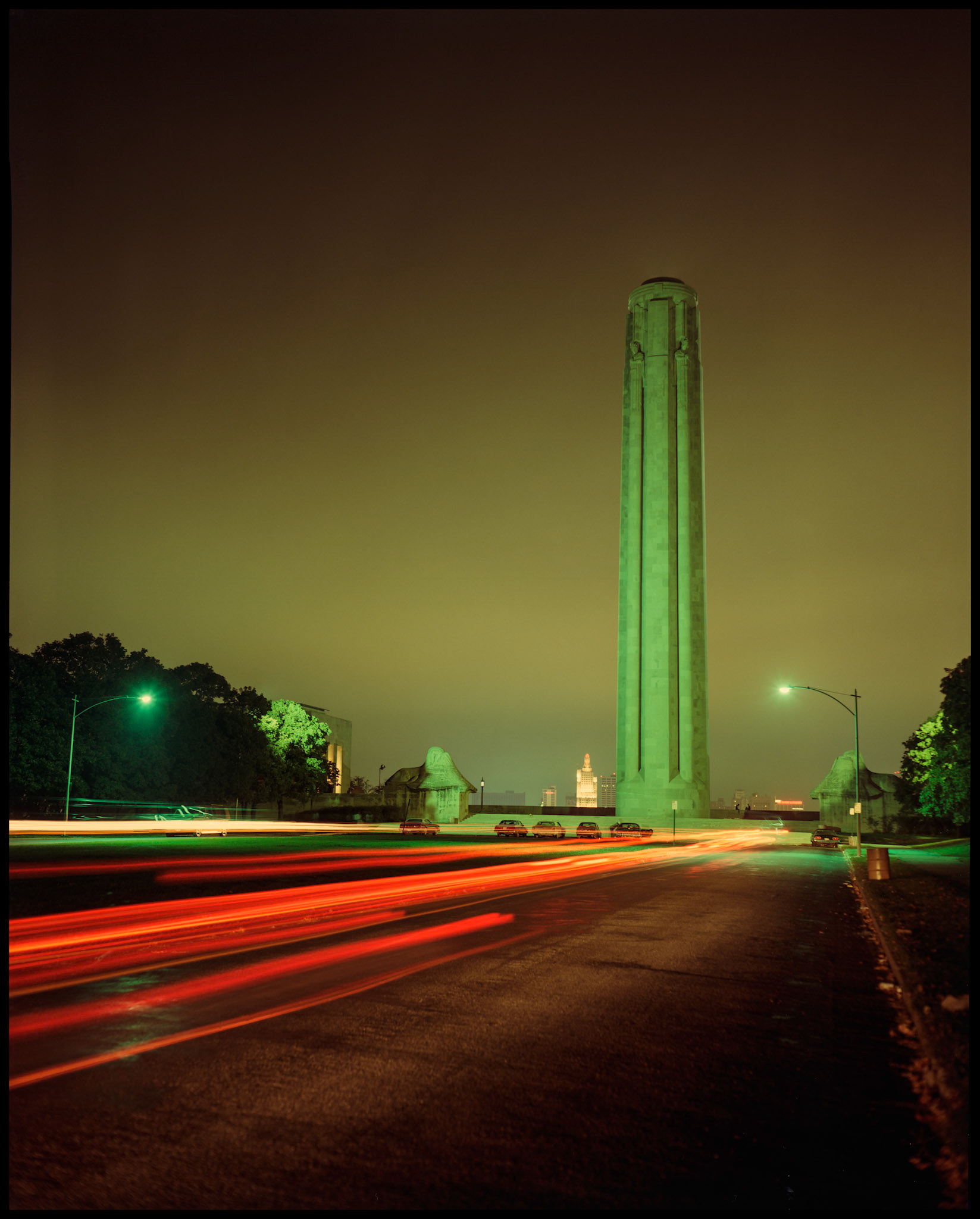 A vintage nighttime time exposure of the Liberty Memorial Tower at the National World War I Museum and Memorial showing streaks of the taillights of passing cars and the Kansas City skyline in the background. Kansas City, Missouri 1984.