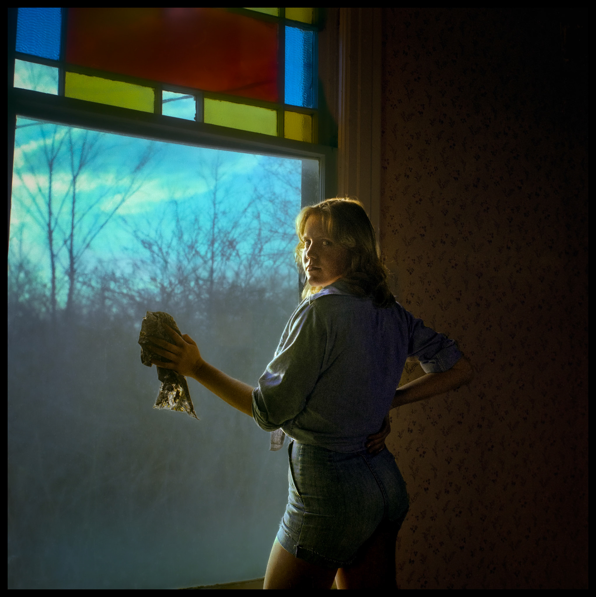 A posed photo of a young woman glancing over her shoulder as she is cleaning the window in an old house. Kirksville, Missouri, 1978