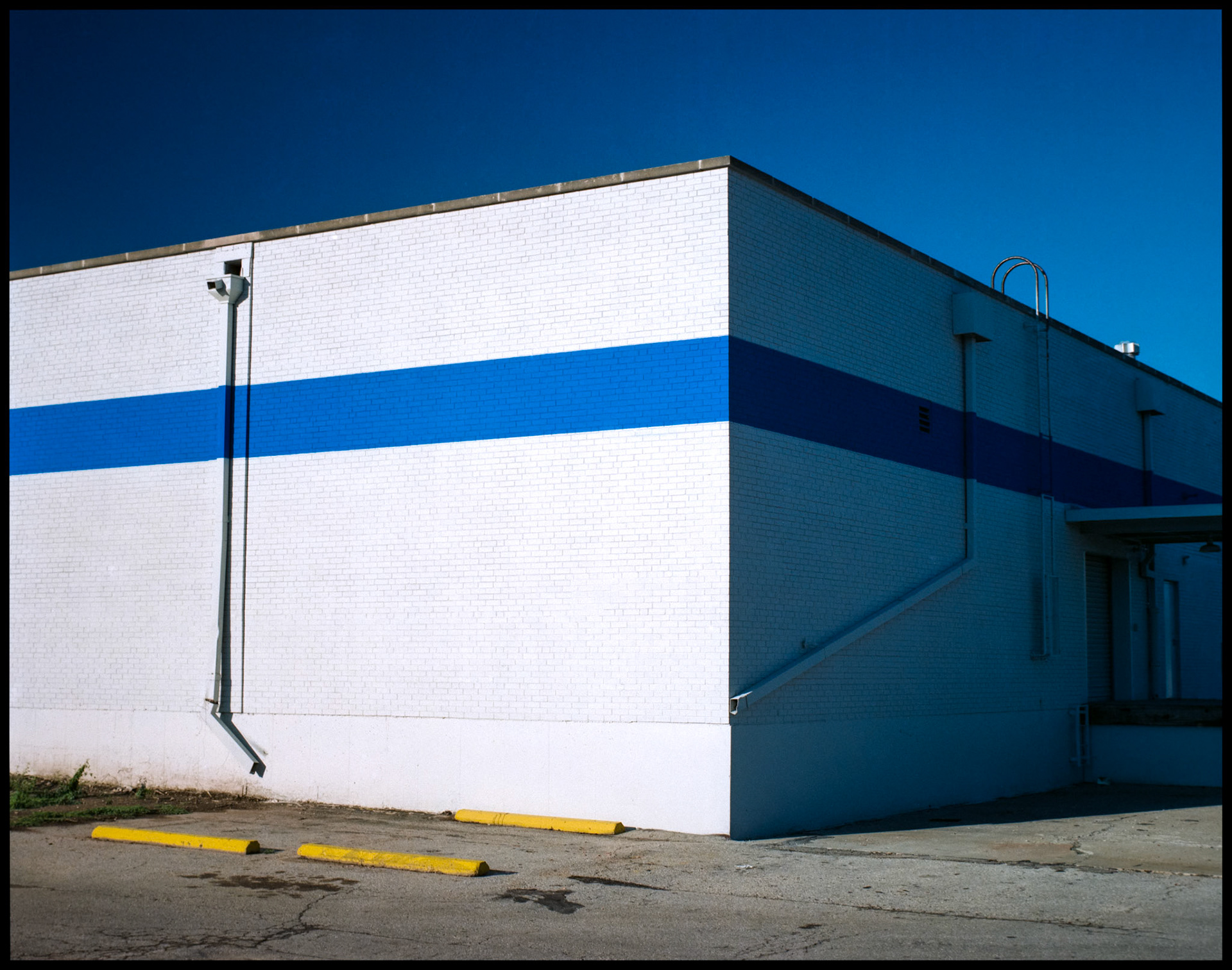 The corner of a brilliantly white building with a blue stripe adorned with drain spouts and an access ladder and yellow parking curbs in the foreground. Columbia, Missouri 1988
