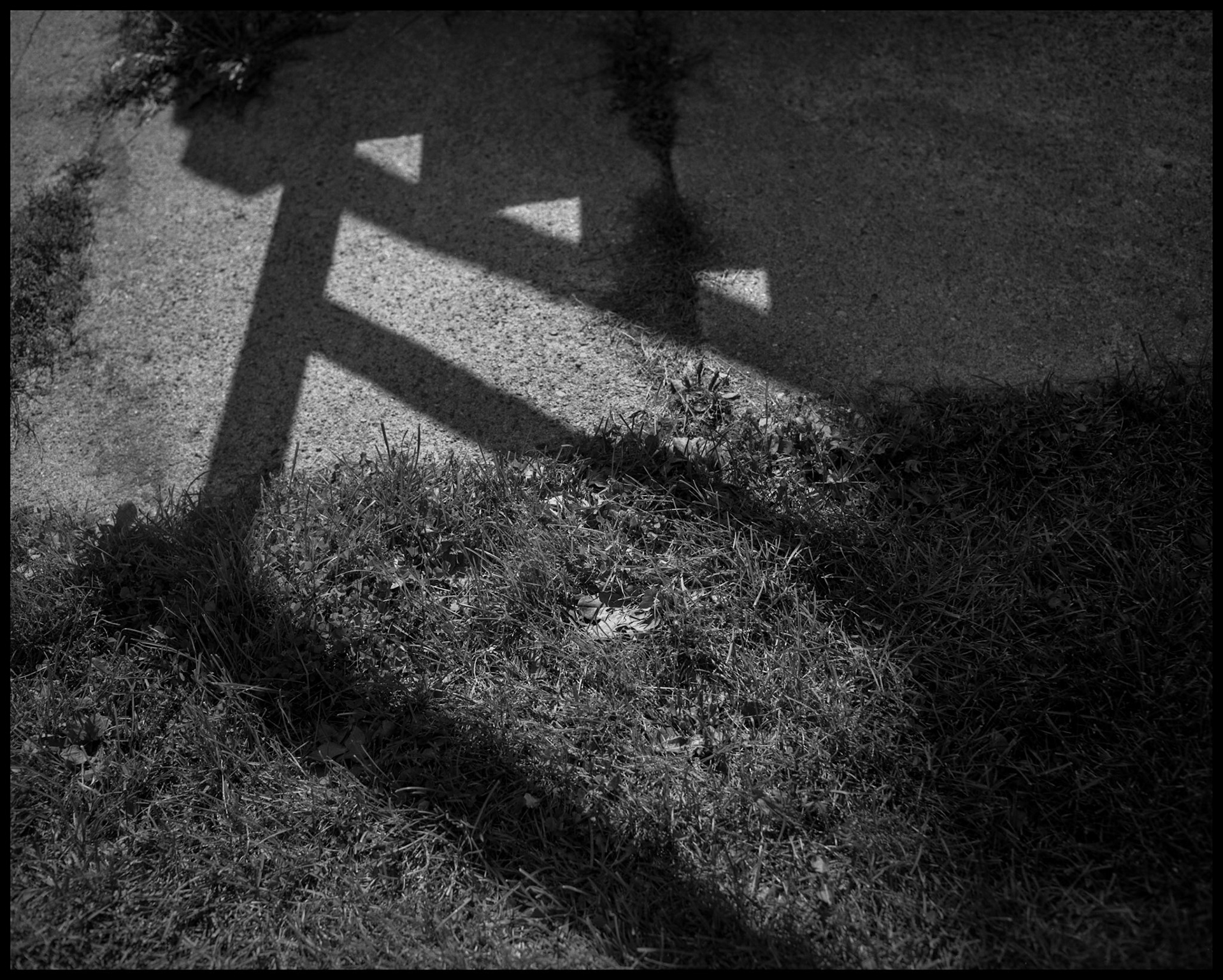 A black and white minimal abstract detail of a leaf in the grass framed by the angular designs created by the shadow of a stair handrail on the sidewalk and grass. Kirksville, Missouri 1984