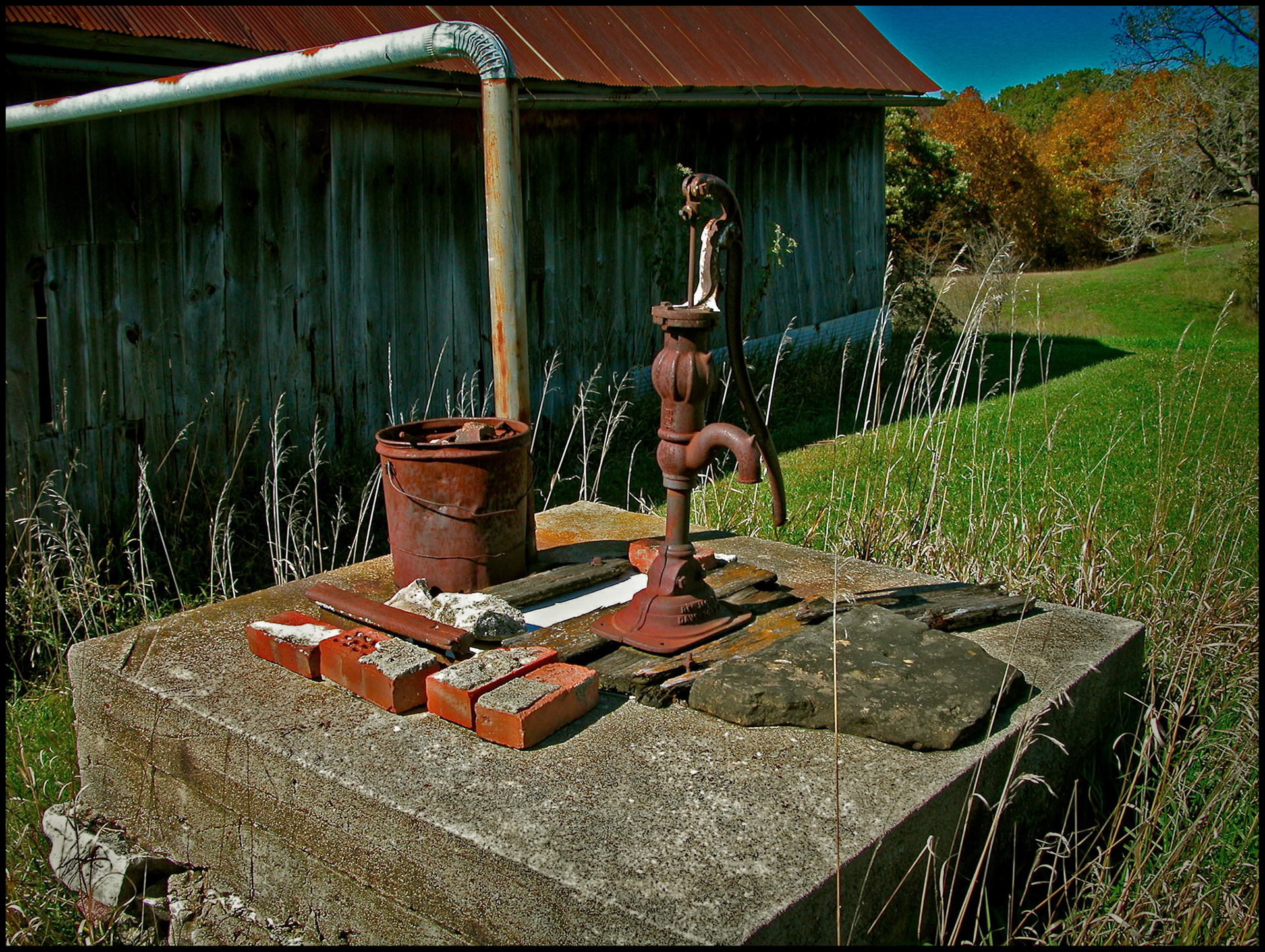 An old well pump on a raised pedestal used to store old bricks, next to an aging barn, with colorful Autumn trees in the background. Near Milan, Missouri, 2003.