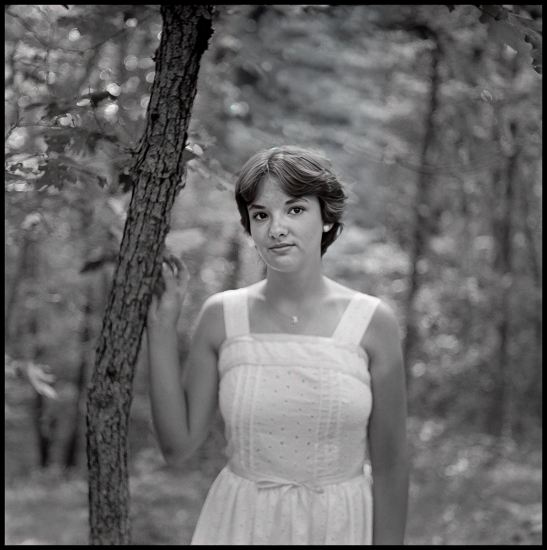 A 1970's vintage soft focus image of a young woman leaning on a tree in a forest with a thoughtful expression. Kirksville, Missouri 1978