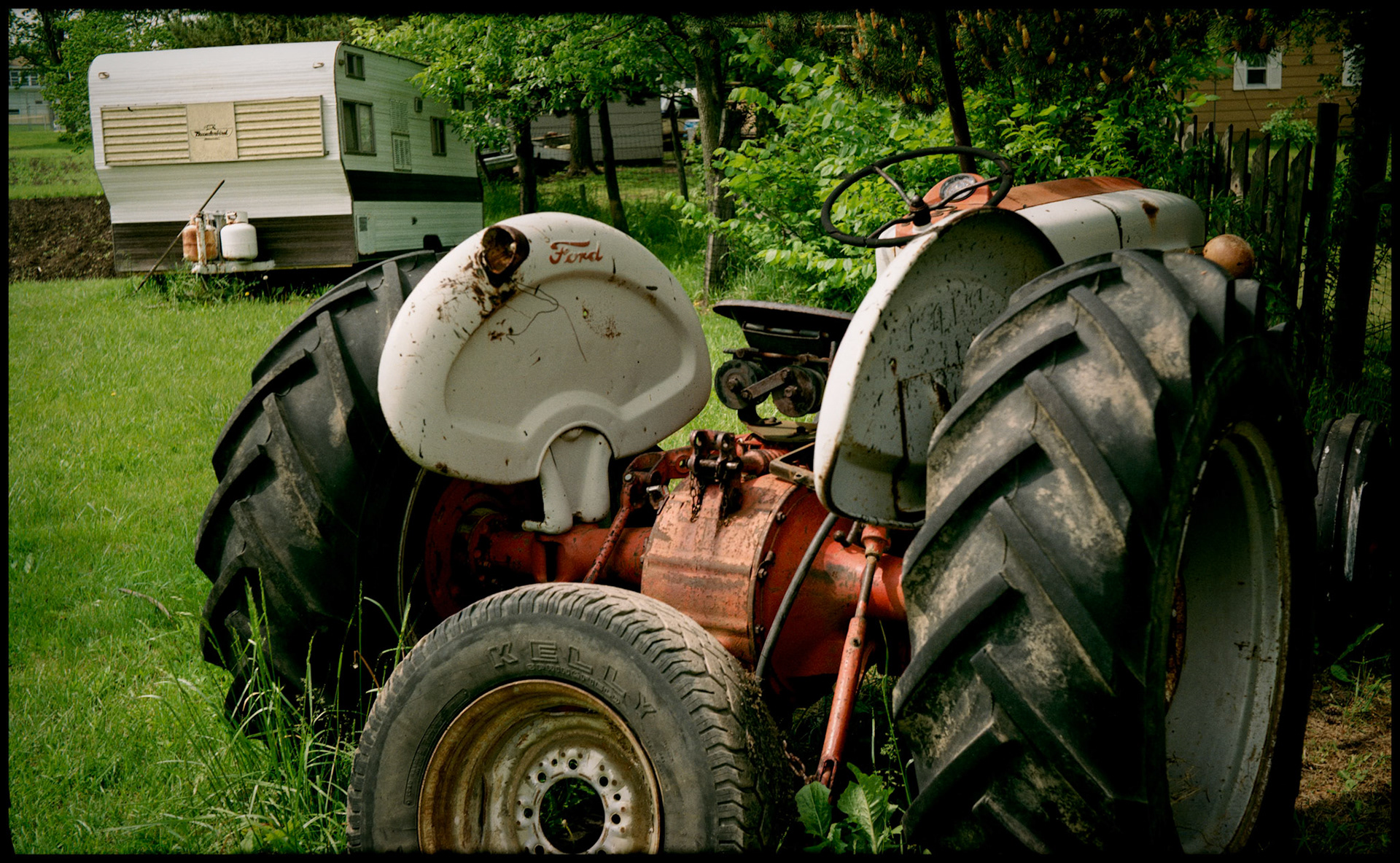 A Ford 8N tractor with a car tire leaning against it and a travel trailer in the background, Green City, Missouri 1990