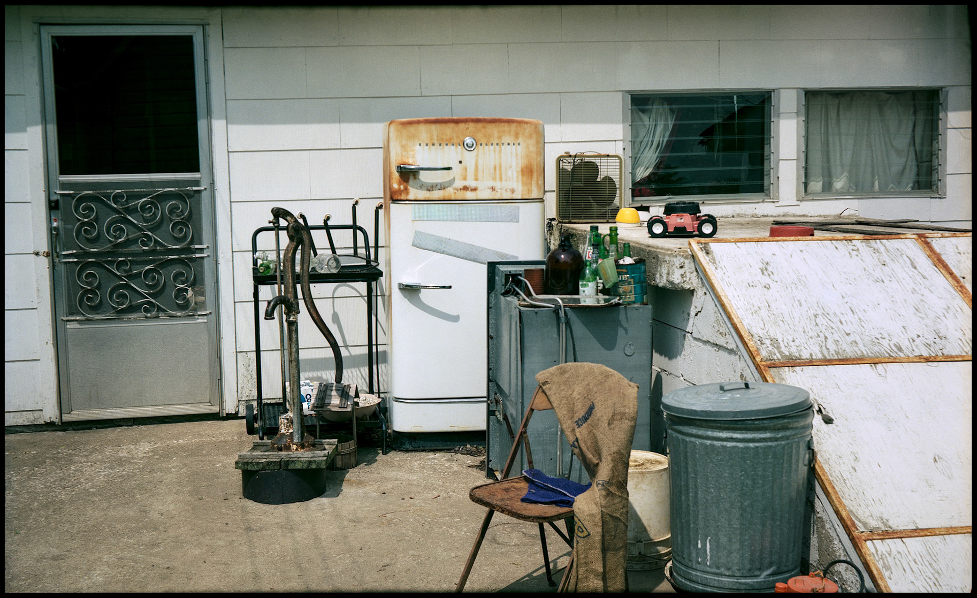 A collection of old appliances and other items surrounding an antique hand well pump on the back patio of a rural small town house.