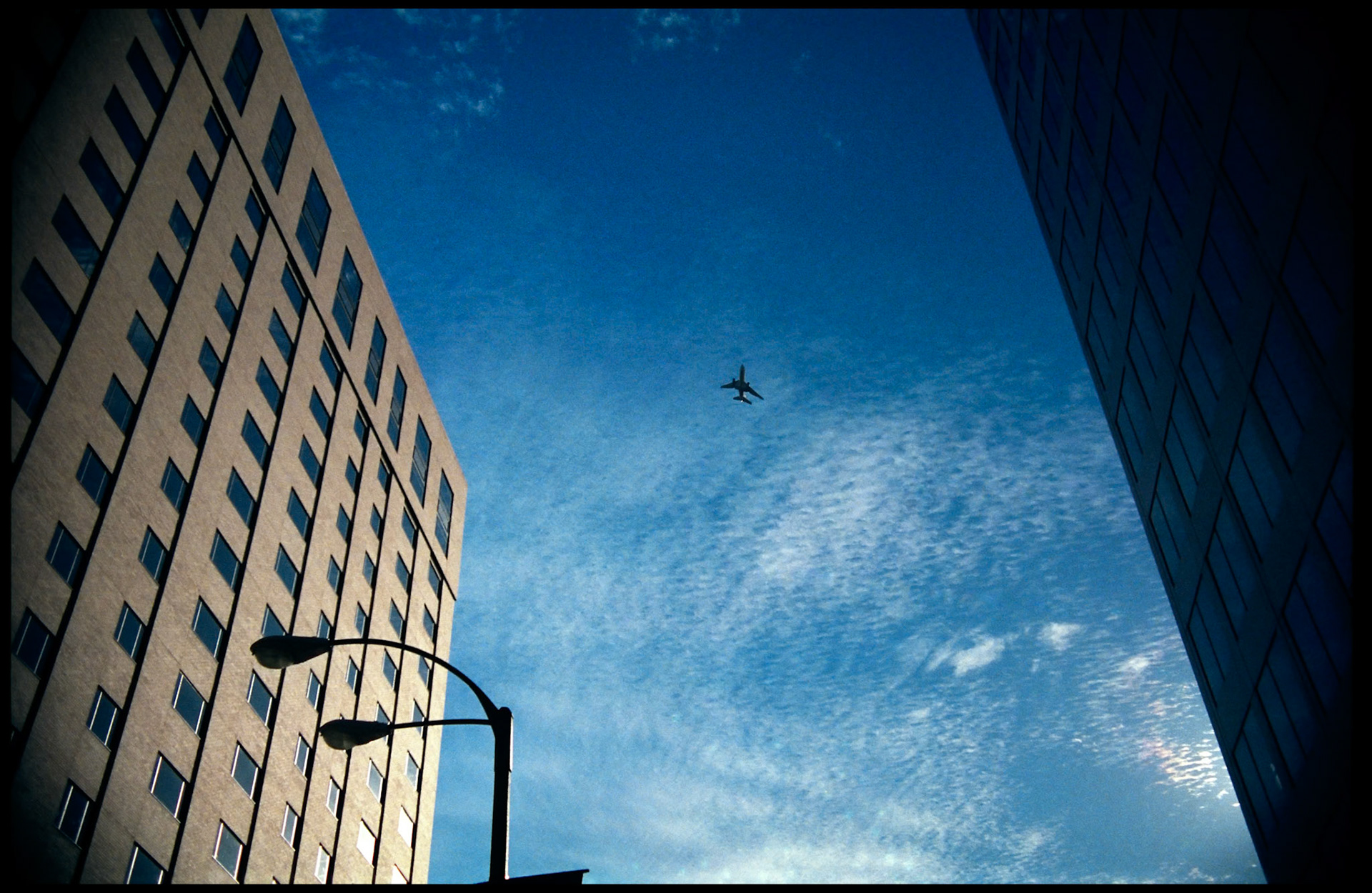 A minimal urbanscape with an approaching airliner framed between two looming builidings and silhouetted streetlamp against a rich deep blue sky with a few textured cirrus clouds. Part of a series shot one warm afternoon in November, 1988 called An Afternoon in St. Louis (a subset of my Industrial Geometry series).
