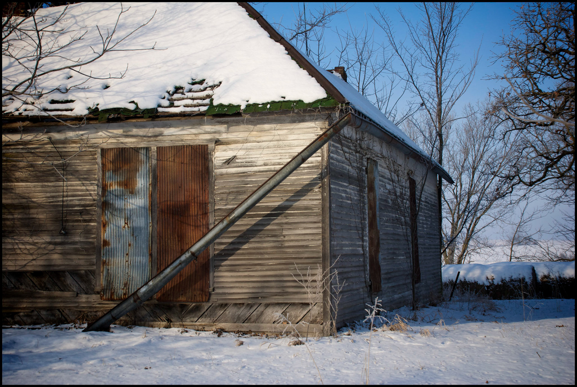 A snowy winter scene of a dilapidated abandoned rural farmhouse with the windows tinned over and a fallen rain gutter surrounded by hay bales and trees in rural Northern Missouri. Near Loeffler, Missouri USA, 2008