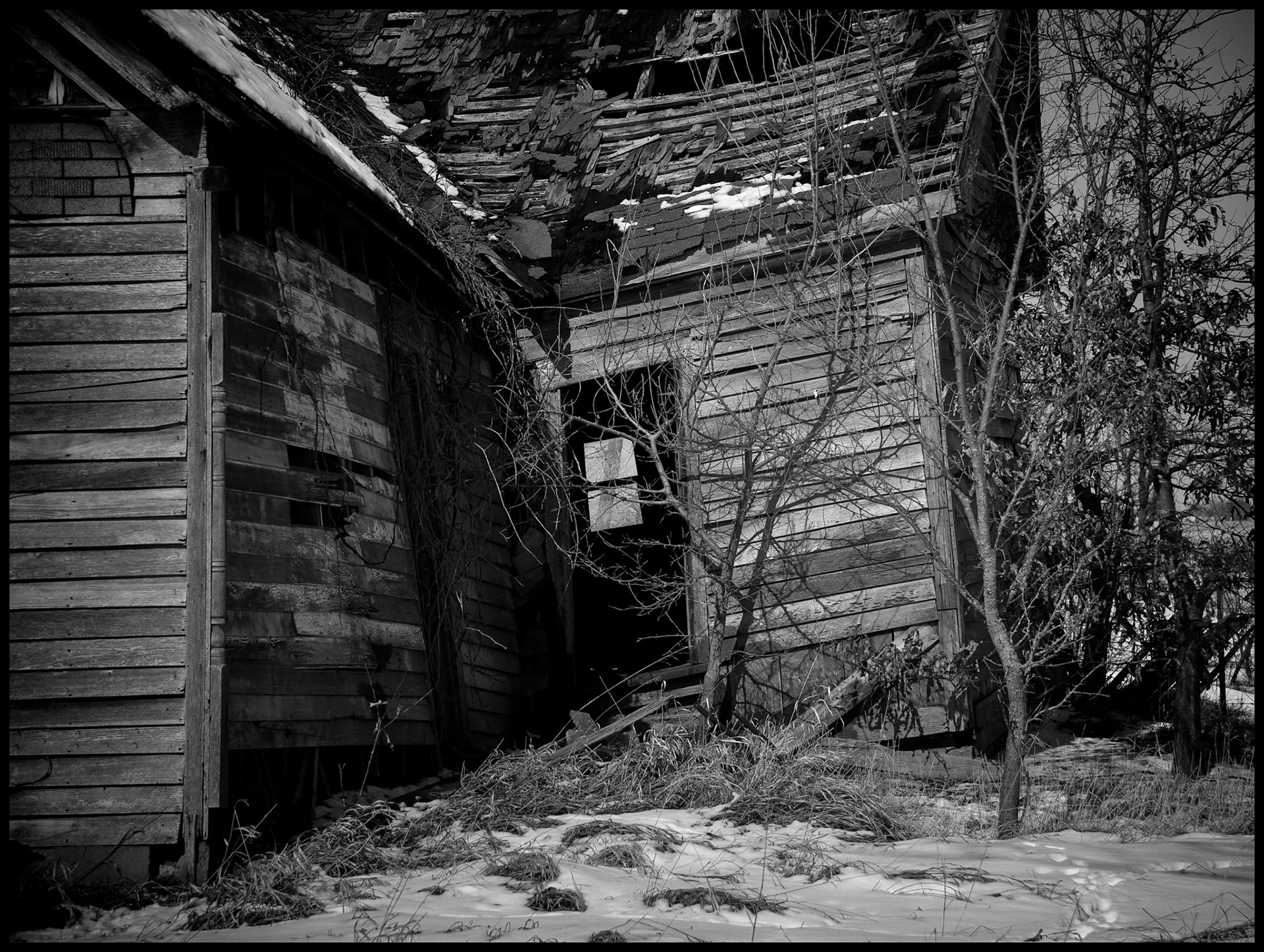A black-and-white rural Missouri winter landscape detail of a dilapidated abandoned house partially covered in snow looking through an open door to a window on the opposite wall. Near Winigan, Missouri USA 2008.