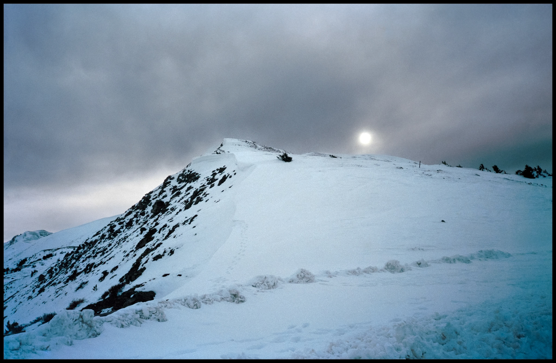 A gloomy snowy day on the top of Cottonwood Pass with the sun peaking through the cloud cover and tracks in the snow. Between Buena Vista and Taylor Park in Southwest Colorado USA 1993