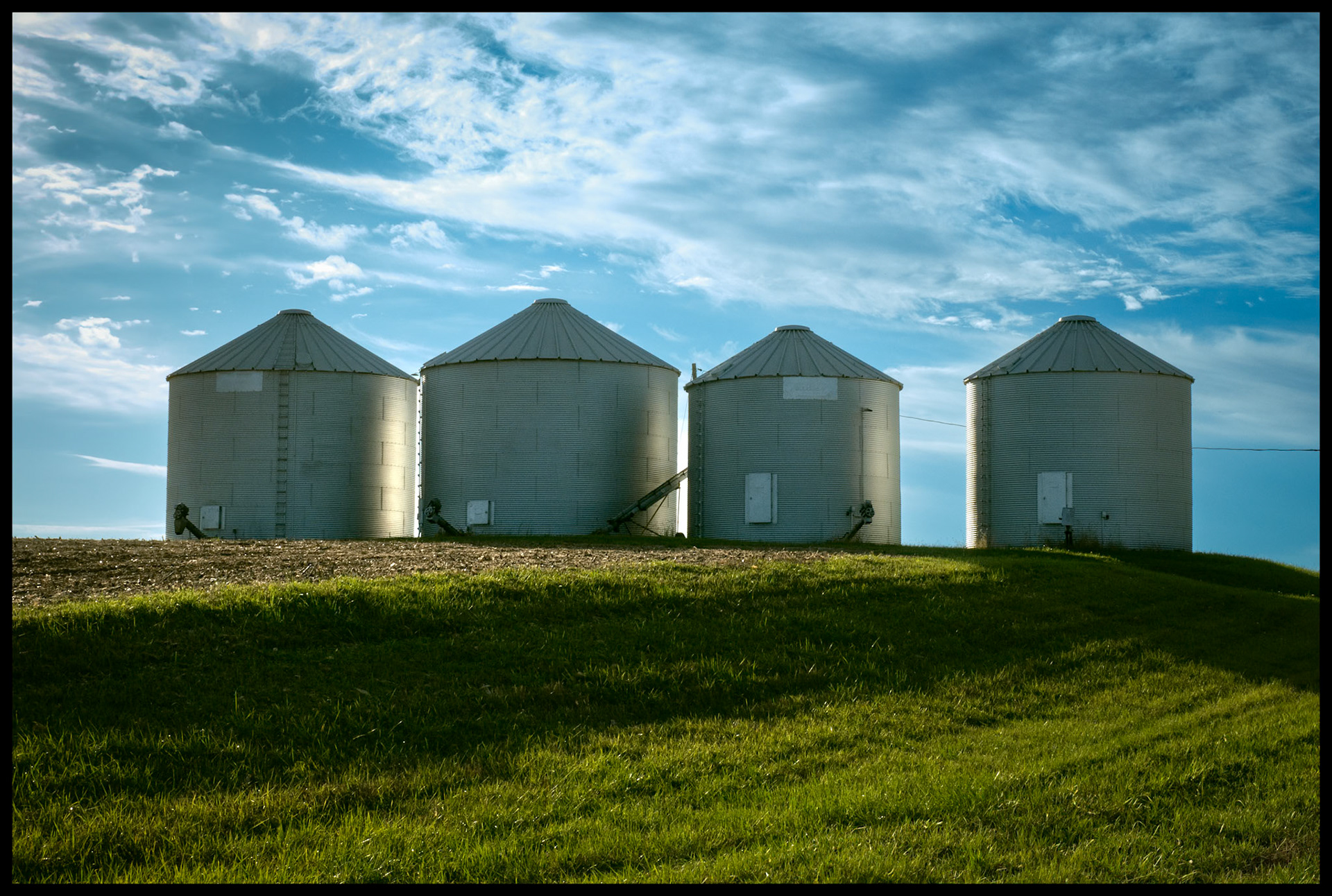 Four backlit grain bins on a hill at the edge of a soybean field right after harvest framed by a blue sky with whispy clouds. Near Ethel, Missouri. 2023