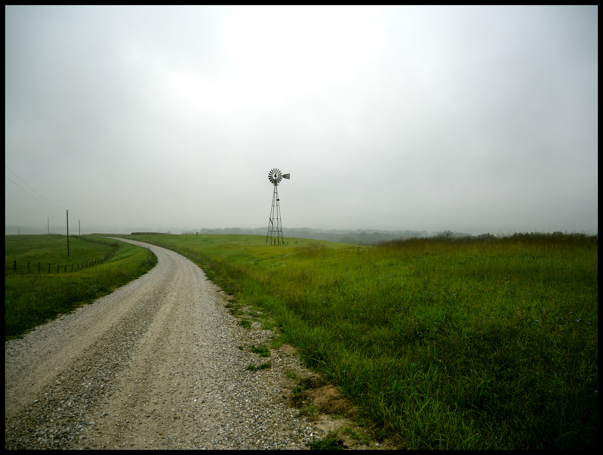 A solitary windmill at the edge of a gravel road and green field silhoutted against the overcast sky on a rainy and foggy September morning. Located on Indian Hill near South Gifford, Missouri, 2023.