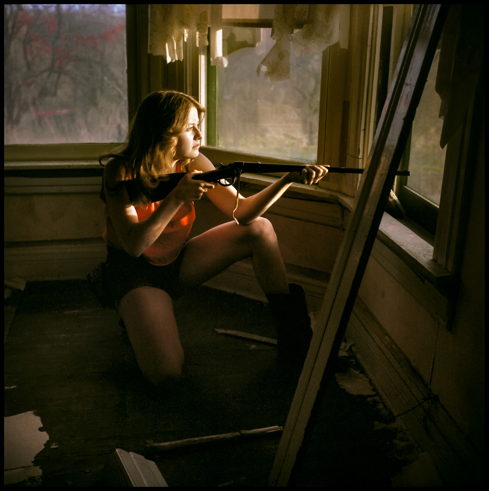 A young woman posed as a hilbilly girl kneeled watching out the window of a house for an imaginary threat with a weapon in hand to defend herself. Kirksville, Missouri. 1978