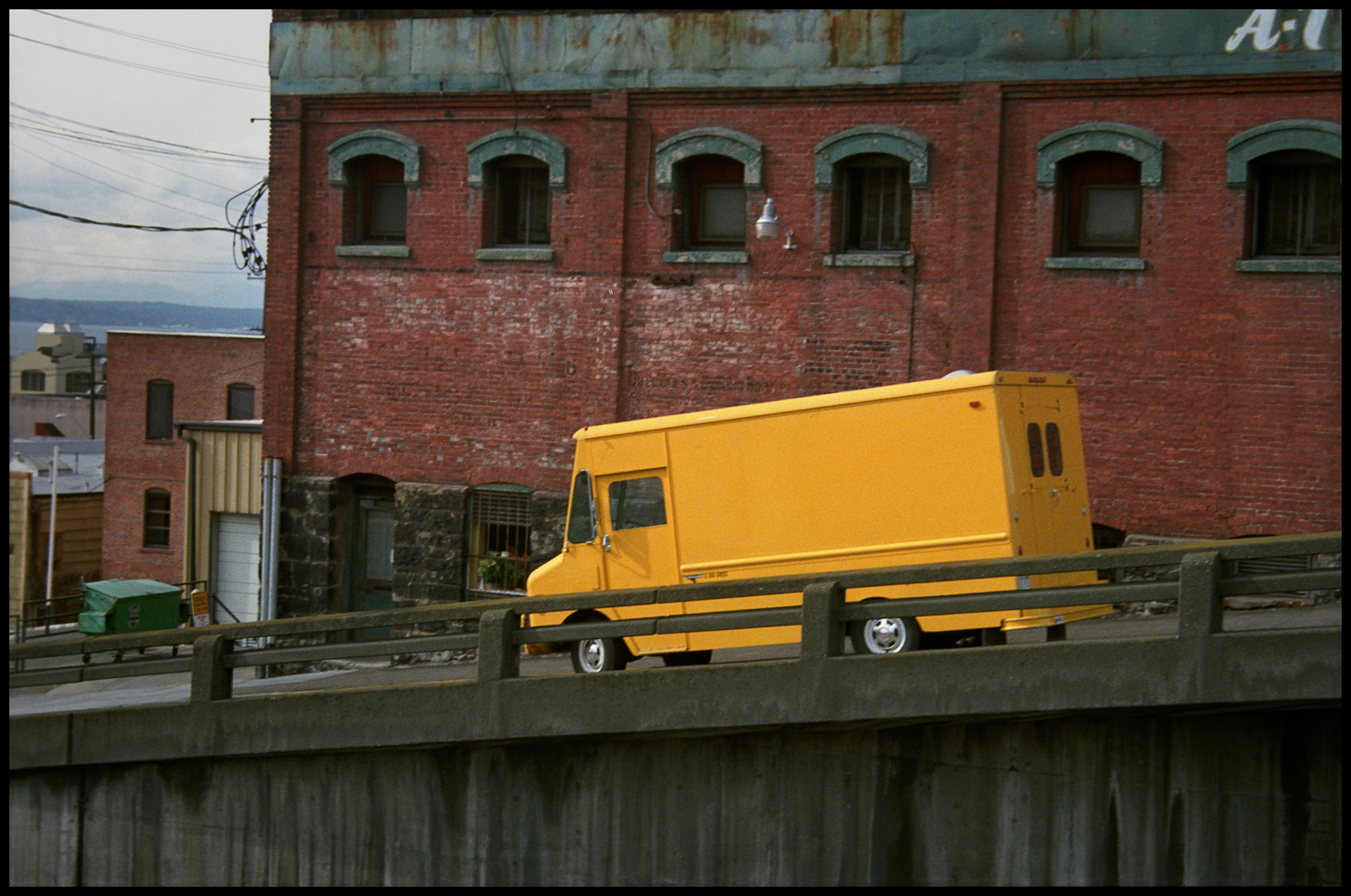 A vintage Seattle Washington street scene of a yellow delivery van parked on a hillside street (First Ave and Battery) in front of a red brick building. 1987