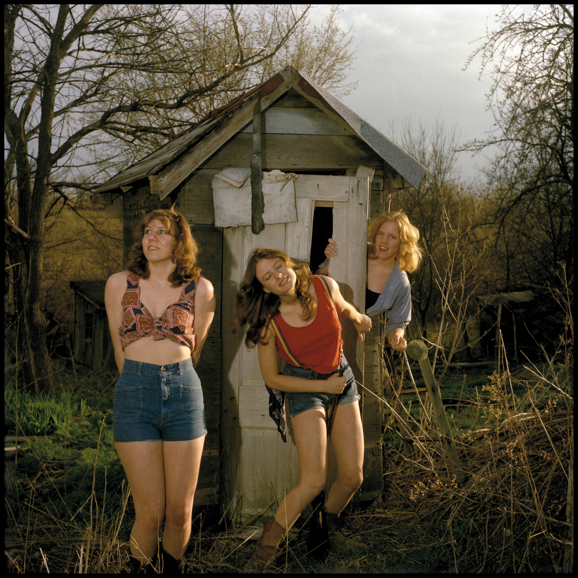 A whimsical vintage image of three teenage girls posing as hillbilly girls with props in front of an abandoned outhouse. Kirksville Missouri 1978
