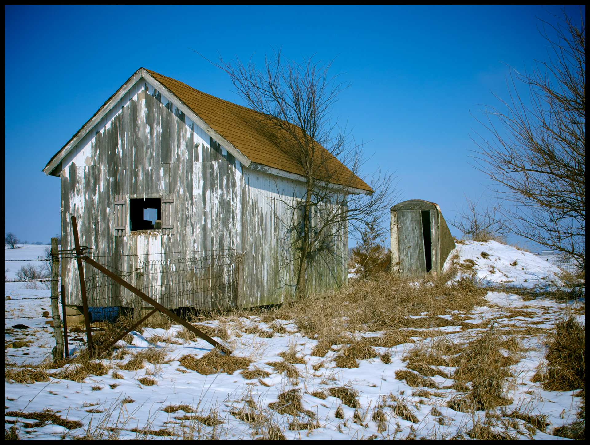 A winter scene of a rural  abandoned shed and storm cellar next to a fence in the snow, near Loeffler, Missouri, 2008