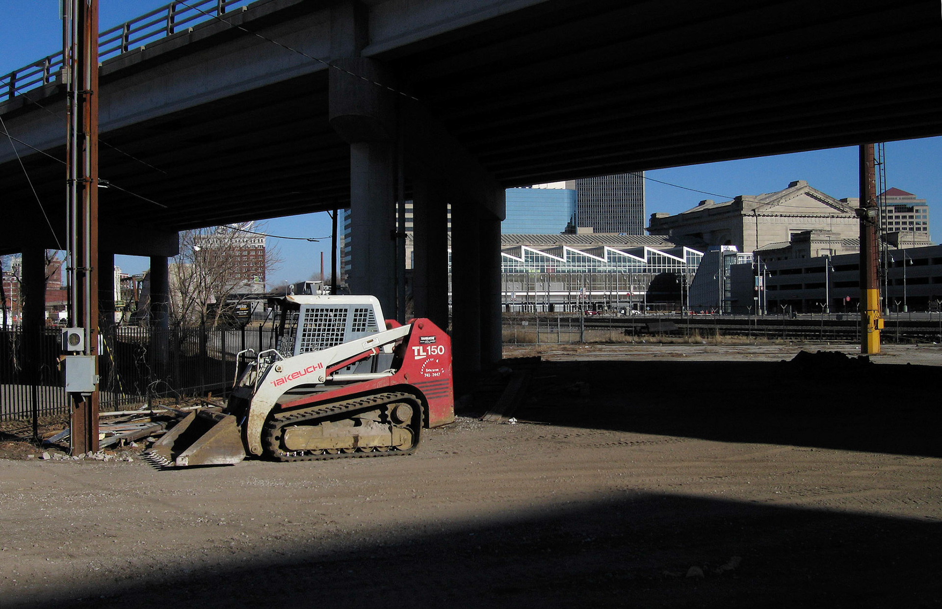 City Urbanscape at the end of Cesar E Chavez drive in Kansas City, Missouri looking underneath the Broadway Boulevard overpass toward the angular roof of the Amtrak Station with a Bobcat type mini excavator in the foreground just beyond a angular dark shadow.