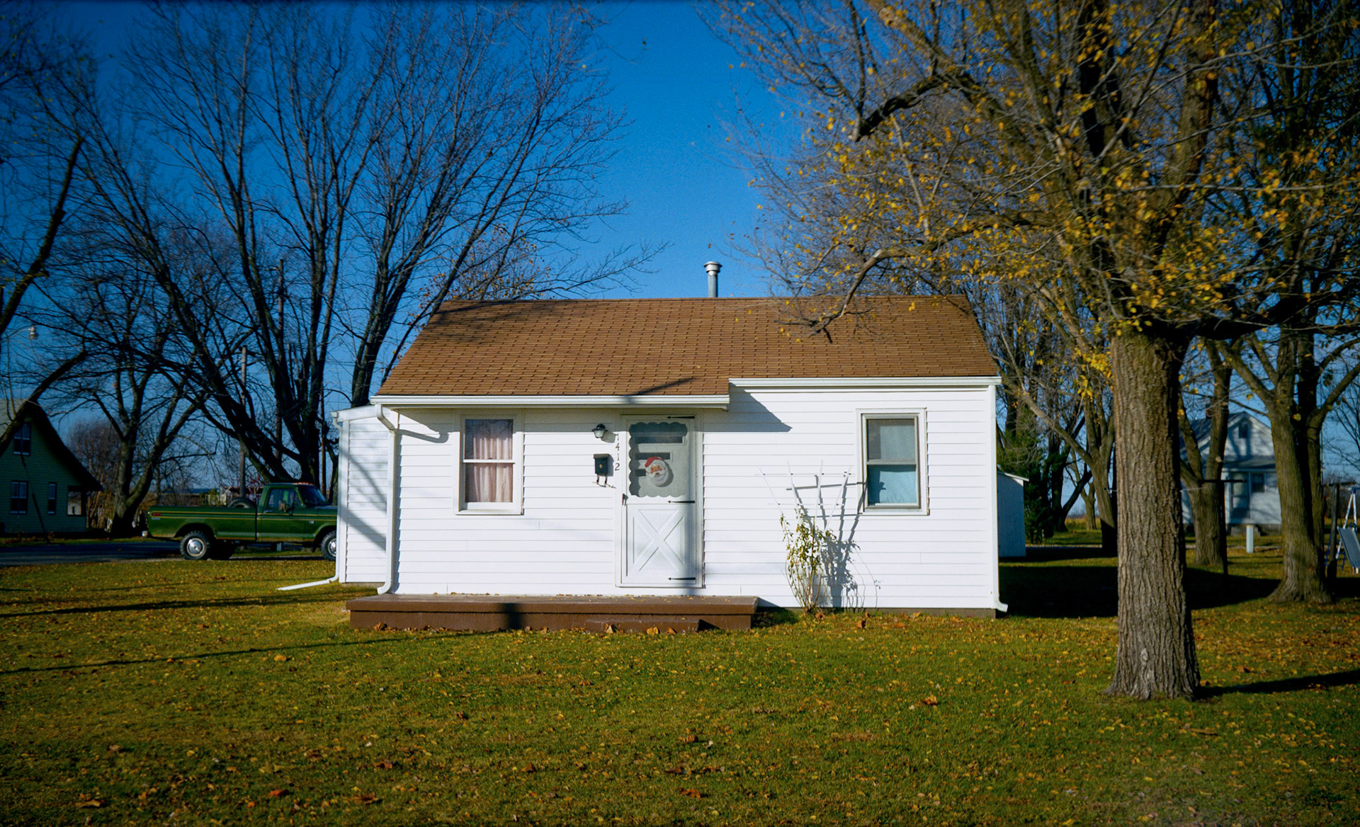 Small white rural house brightly lit by autumn sun with a Santa Claus figure in the front door window.