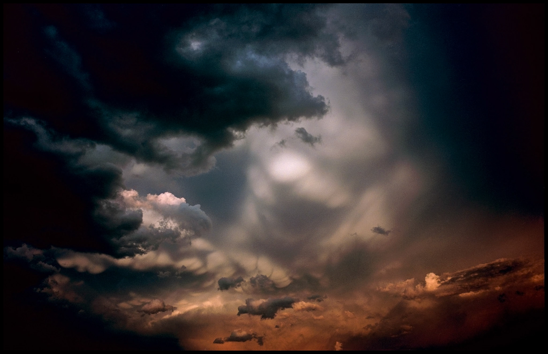 Dramatic turbulent clouds from an approaching storm front brightly lit by the warm light of the setting sun through an opening in the clouds producing a shape resembling a man's face getting ready to blow. My front yard near Renick, Missouri, 1996.