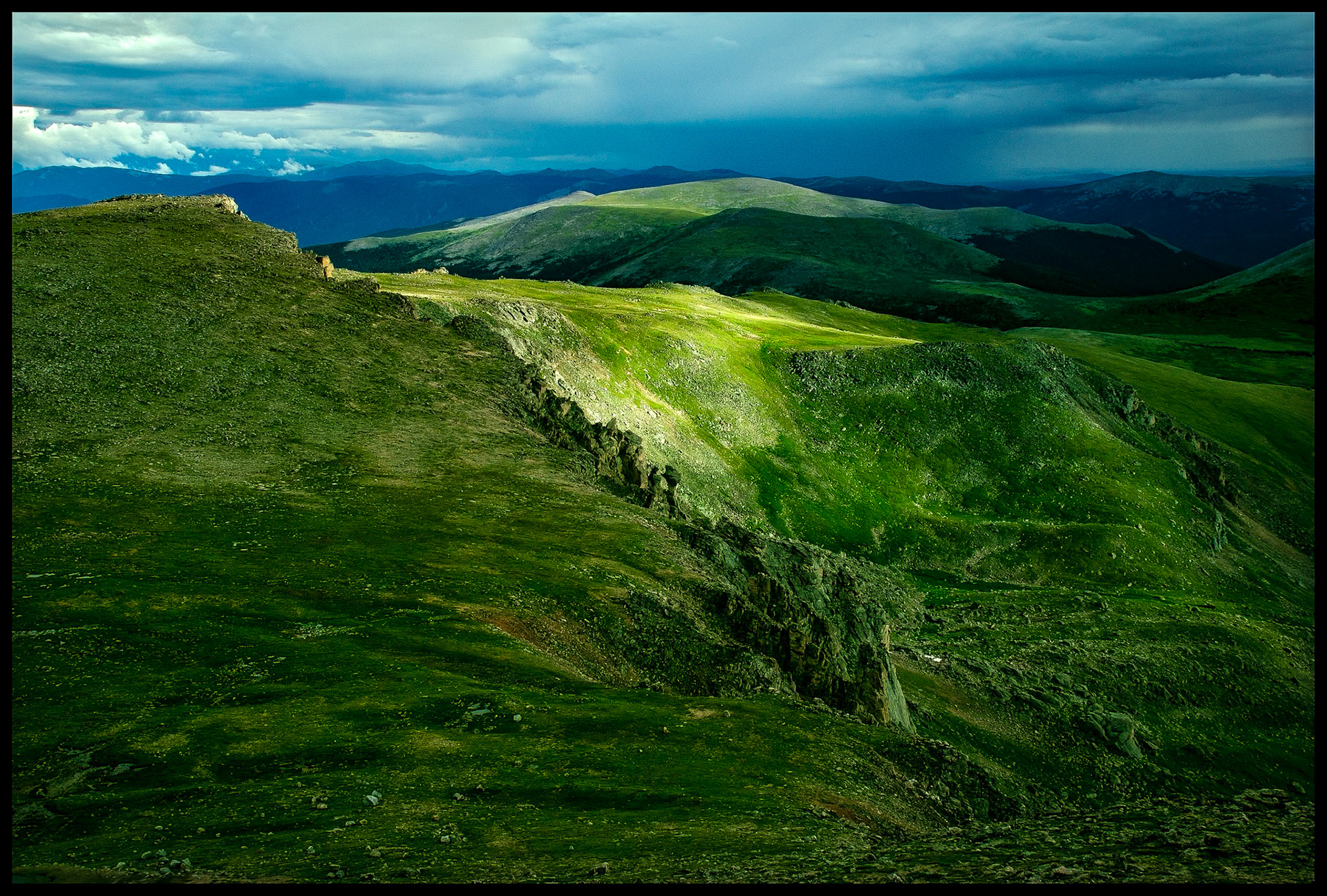 A spot of sunlight on a  mountain ridge highlighting the green rocky tundra with a panoramic view of the surrounding mountains in the background. View from Mount BLue Sky Scenic Byway, near Idaho Sprigs, Colorado USA, the highest paved road in the country.