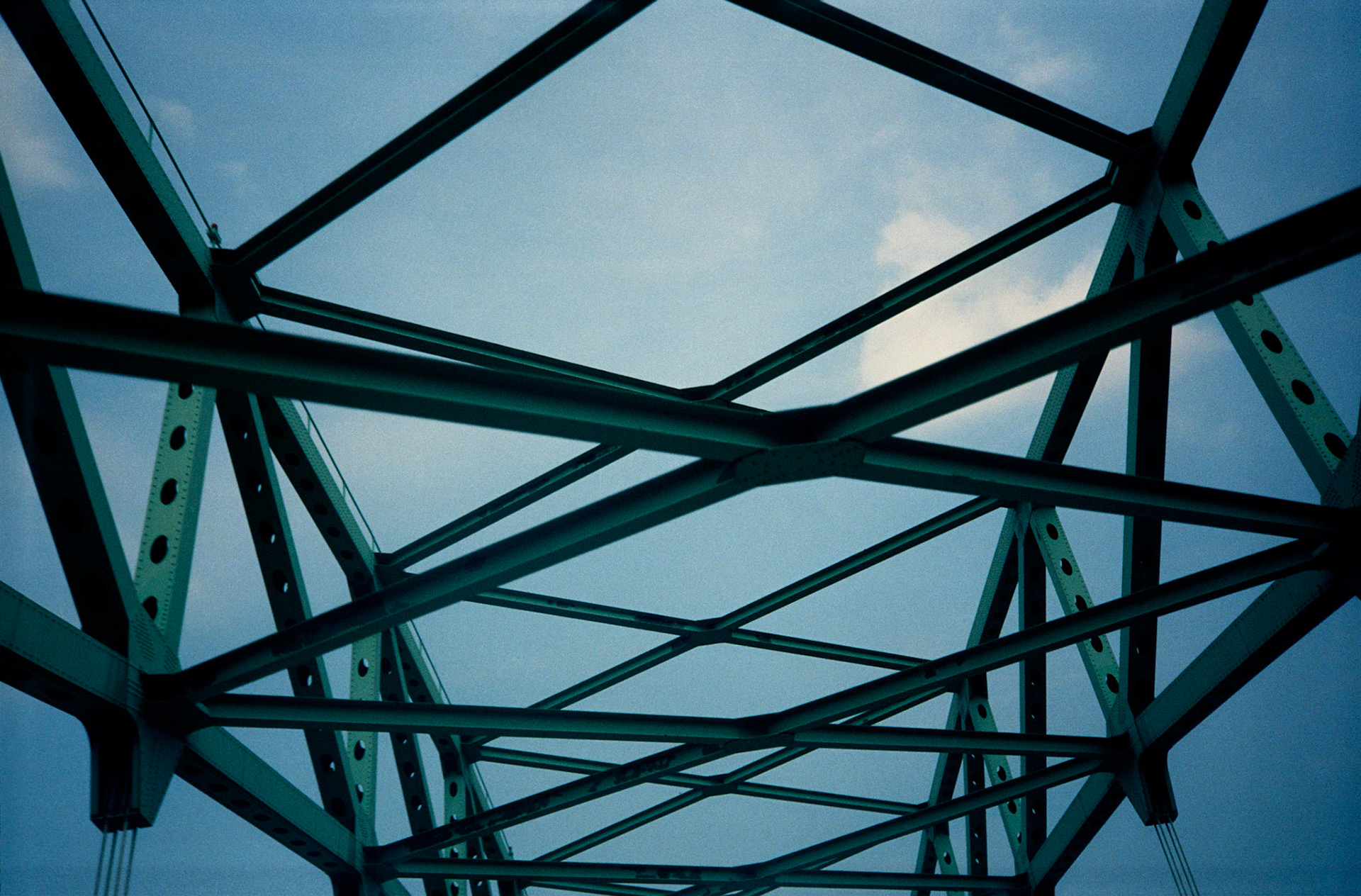 An upward view of the angular patterns created by green iron bridge trusses against a pale blue sky. Kansas City, Missouri, 1991
