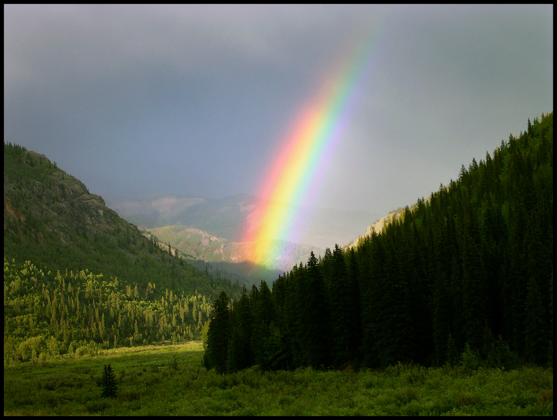 A brightly sunlit rainbow emerging from the mist of a rain shower at the end of a Colorado Rocky Mountain valley between the base of two mountains. Alpine Loop Road near Lake City, Colorado 2004
