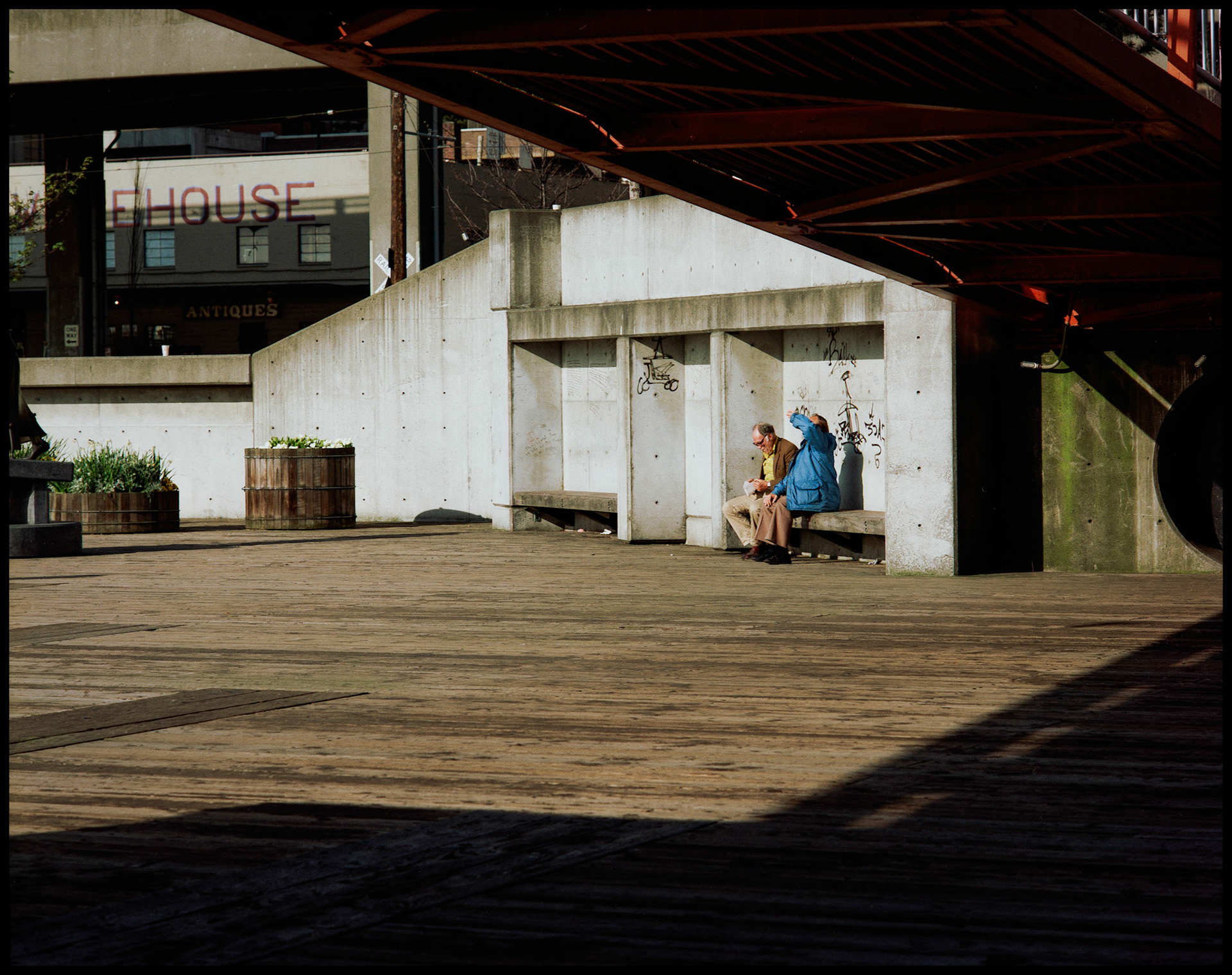 A seattle Washington street scene of two men, one in a blue coat, one in a yellow shirt, sitting on a park bench framed by a walkway and it's shadow. Seattle, Washington, USA, 1988