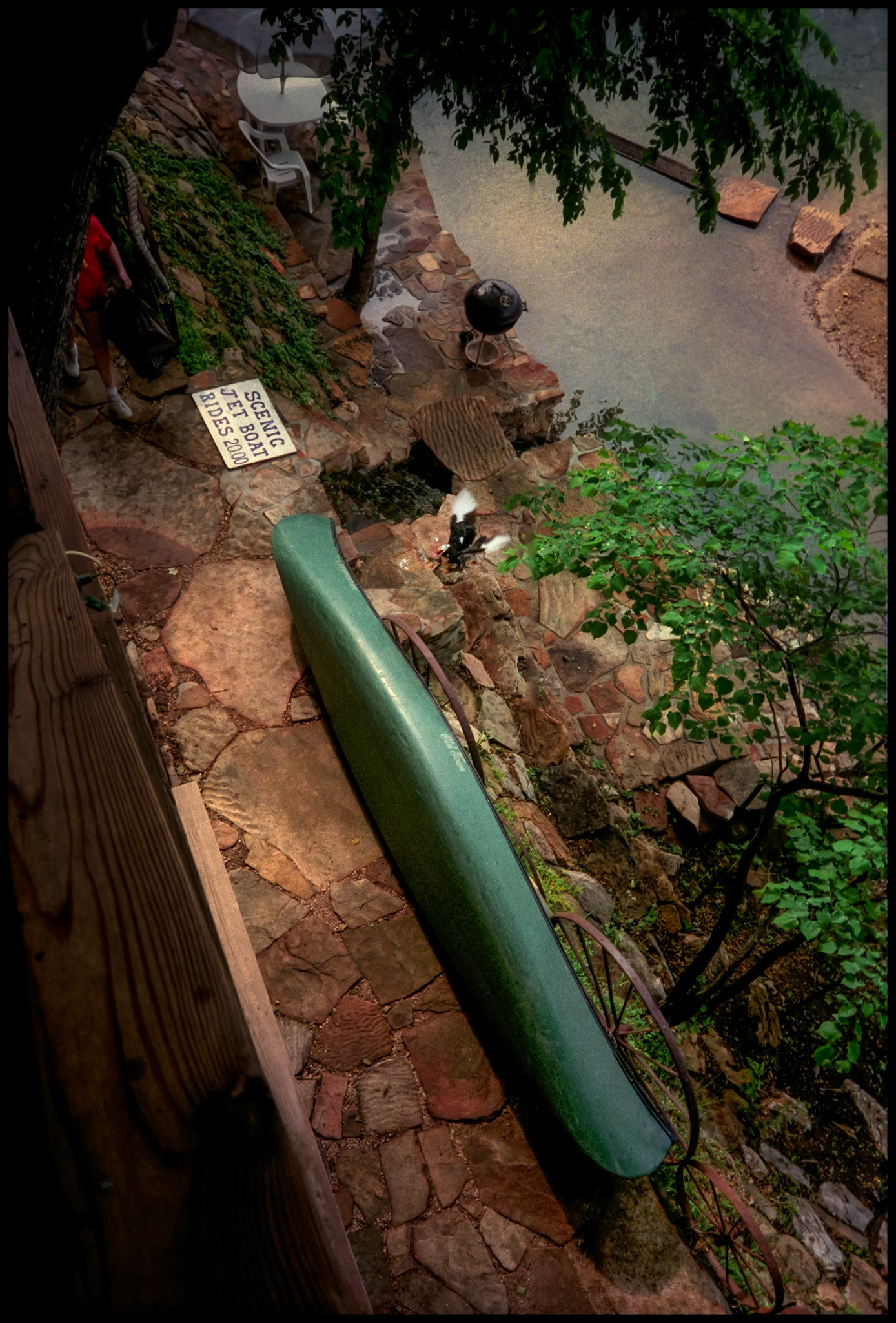 View of a canoe laying on it's side, a duck flapping it's wings, and a comical sign advertising "Scenic Jet Boat Rides". Shot from the balcony of Rivers Edge Resort, Eminence, Missouri at the edge of the Jacks Fork of the Current river in 1992.