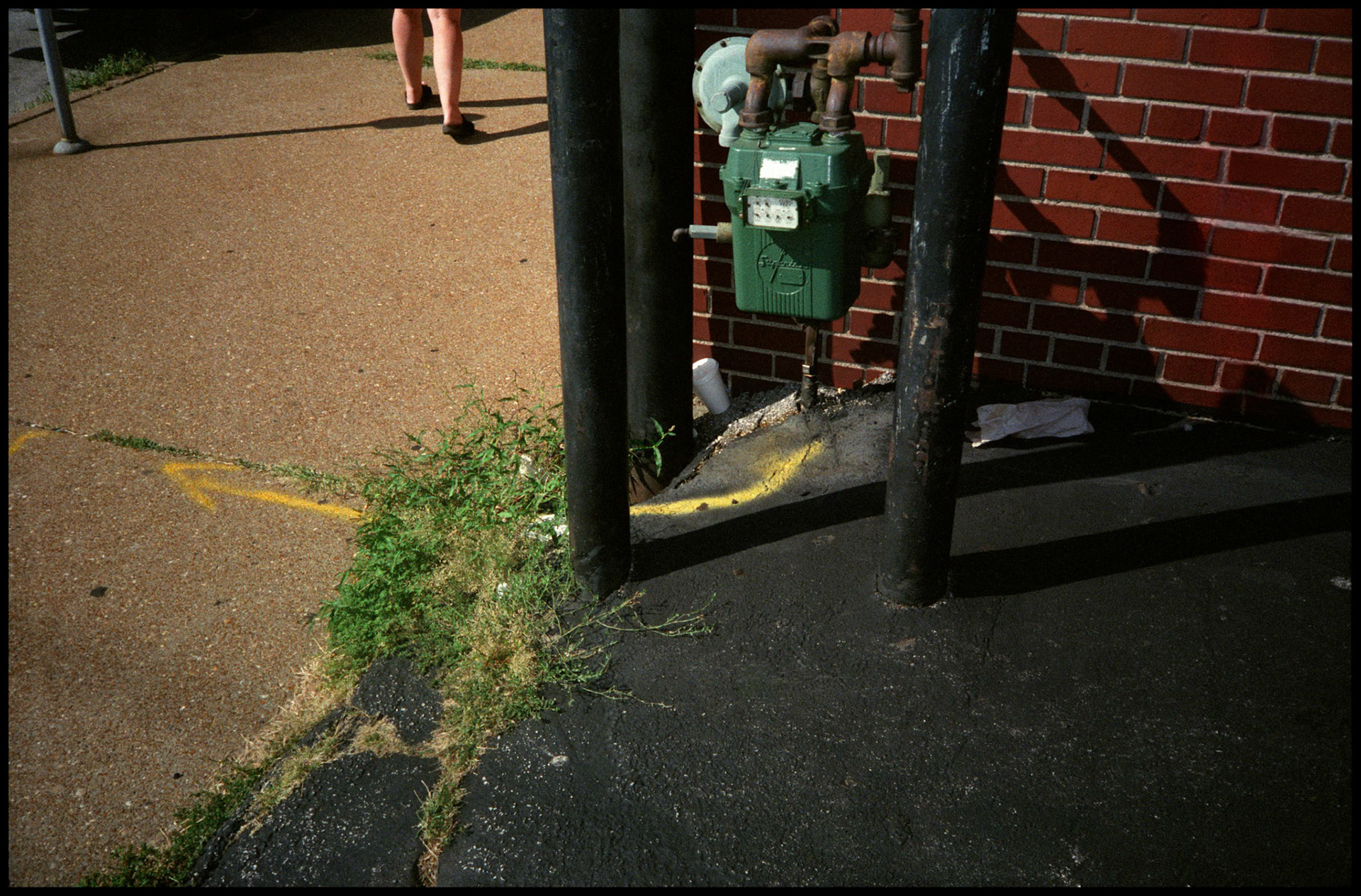 Conceptual abstract detail image of a Natural Gas meter  next to a brick building with safety barrier poles around it and a painted arrow extendning away from it onto a sidewalk. Also on the sidewalk leading out of the frame are a woman's legs. St. Louis, Missouri 1991
