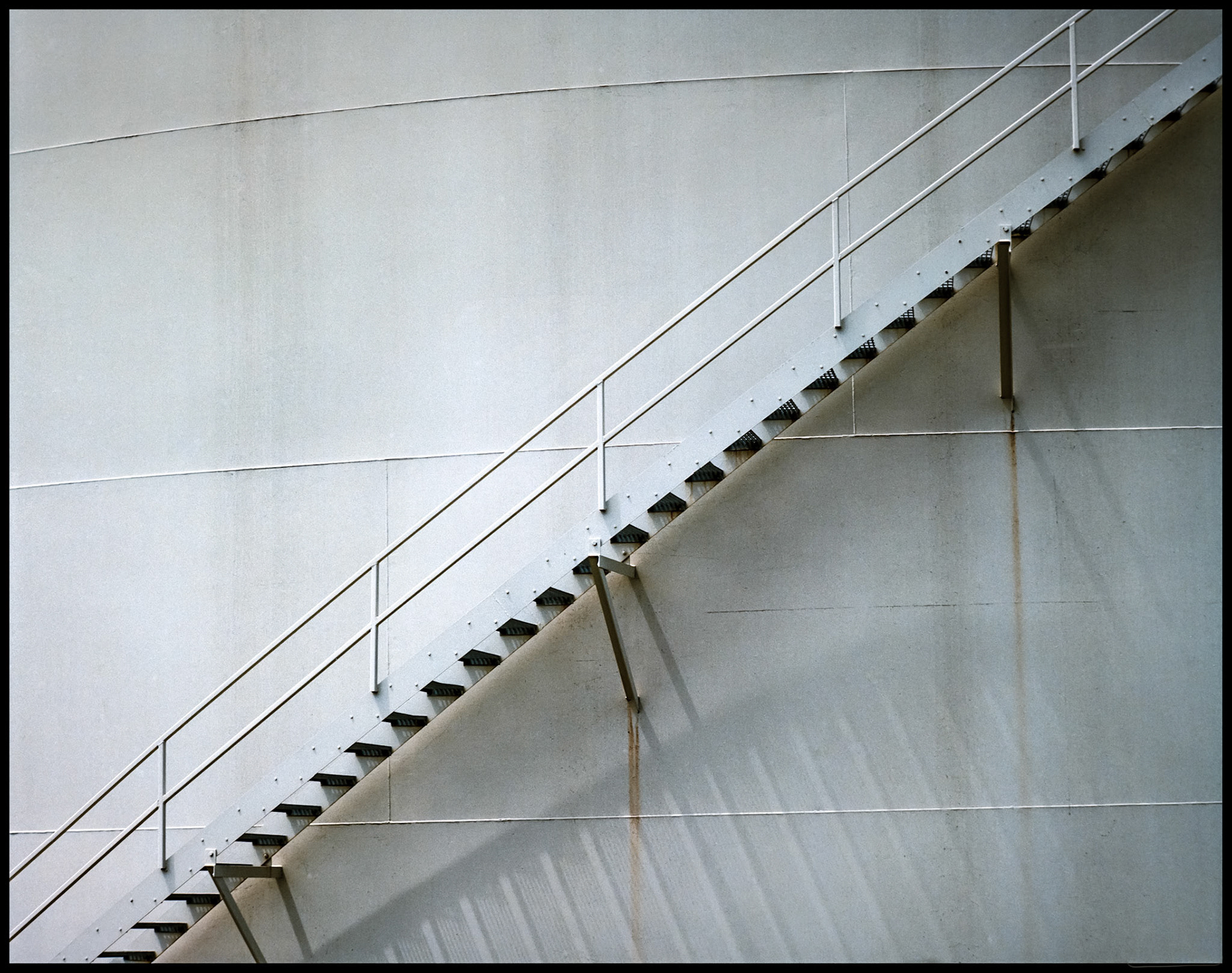Detail of the stairway leading up the side of a large fuel storage tank with rust stains and interesting shadows from the stairs at the Magellan Pipeline facility in Columbia, Missouri in 1988.