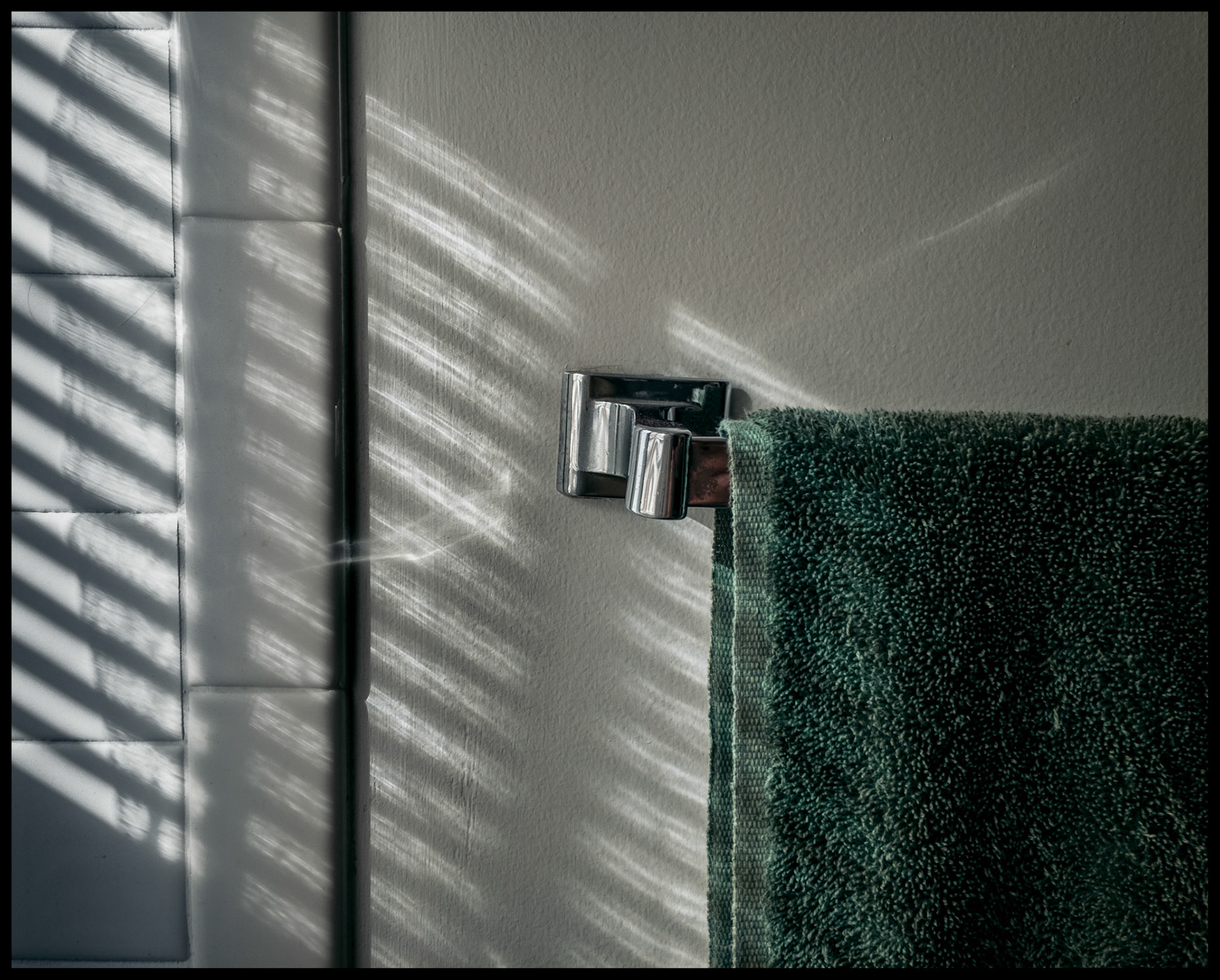 A minimal abstract still life view of highlights and shadows from a window blind on a bathroom wall and towel rack. Kirksville, Missouri 2022
