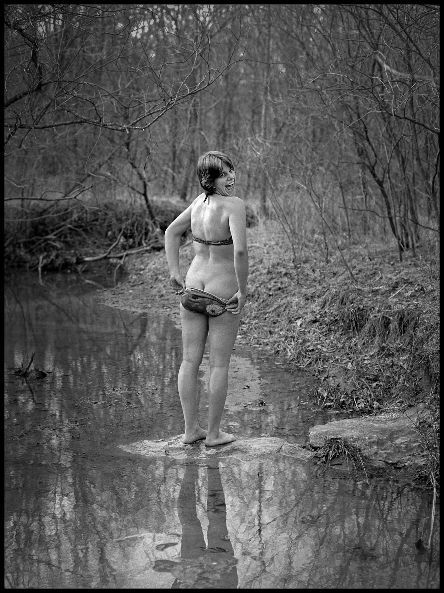 A 1980's vintage image of a young woman in a bikini standing on a rock in a creek playfully teasing a reveal. Kirksville, Missouri 1980