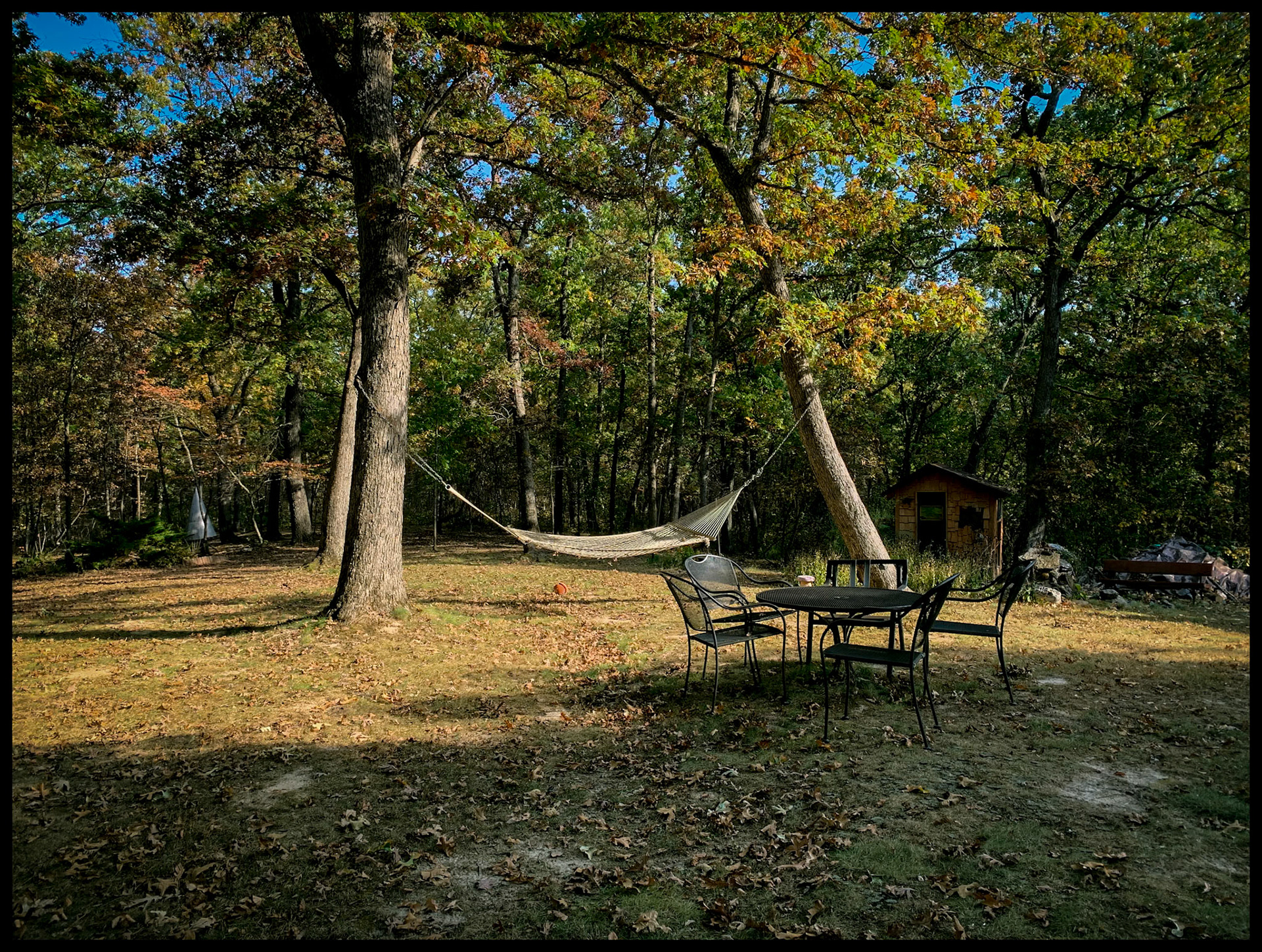 A hammock supended between two trees in a  forested backyard with a table and chairs in the foreground during the leaf color change of Autumn.