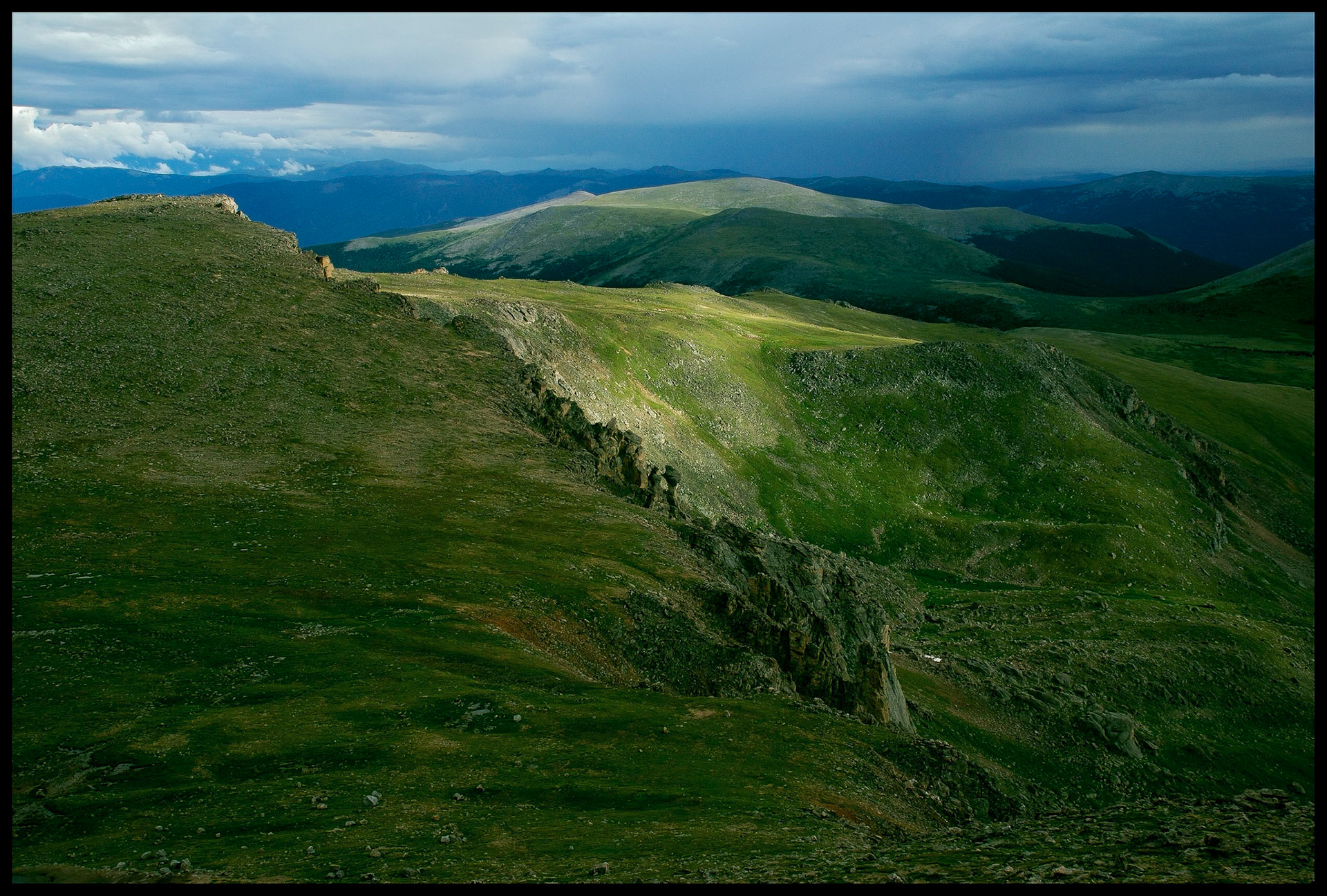 A spot of sunlight on a  mountain ridge highlighting the green rocky tundra with a panoramic view of the surrounding mountains in the background. View from Mt. Evans Highway Colorado USA, the highest paved road in the country.