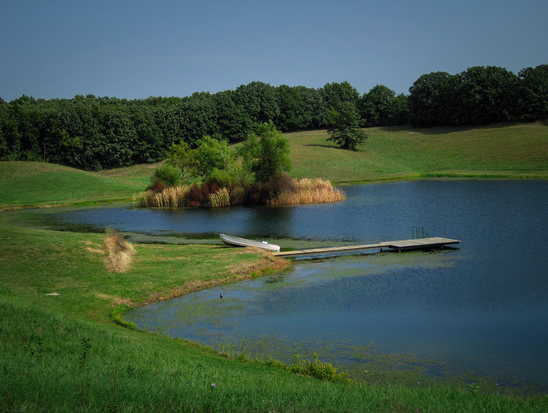 An idyllic scene of a boat on shore next to a small dock on a rural lake with an island surrounded by cattails. Near Yarrow, Missouri USA, 2005
