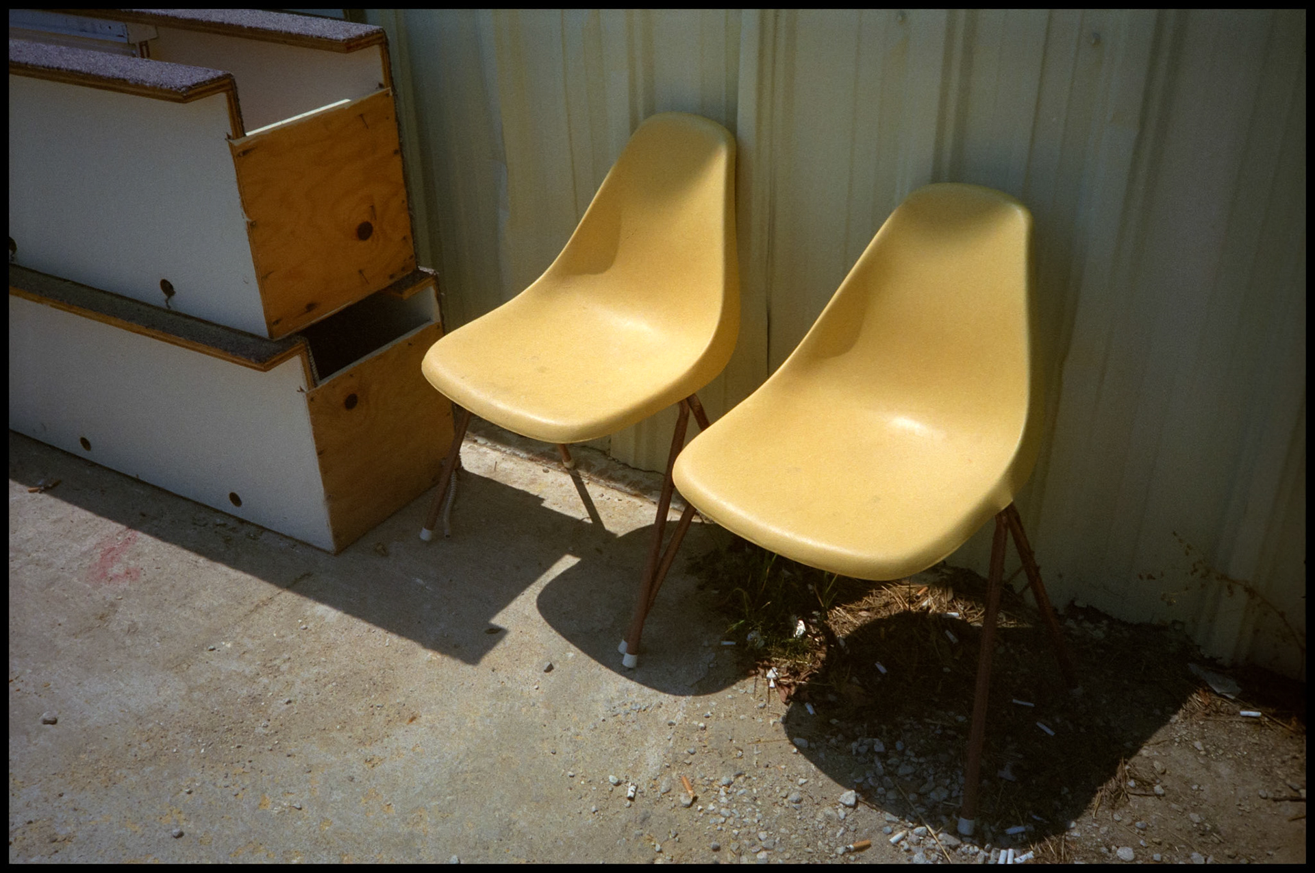A pair of retro yellow plastic chairs sitting outside on the concrete next to a metal building and some wooden boxes.