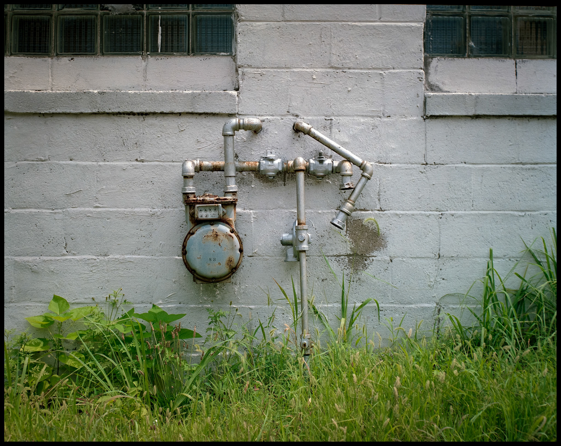 A silver steel gas meter against the side of a white concrete block building in Columbia, Missouri in 1988 with overgrown green grass and weeds in the foreground.