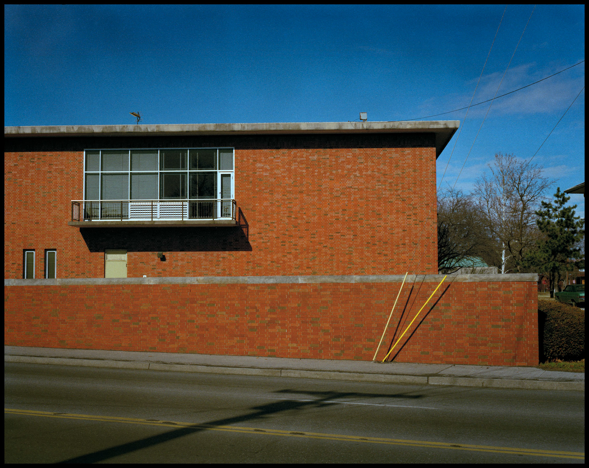 An urban street scene of a red brick building and wall with uitility pole guy wires in front of wall and shadow of another power pole and transformer seemingly pointing toward it. Columbia Missouri 1990