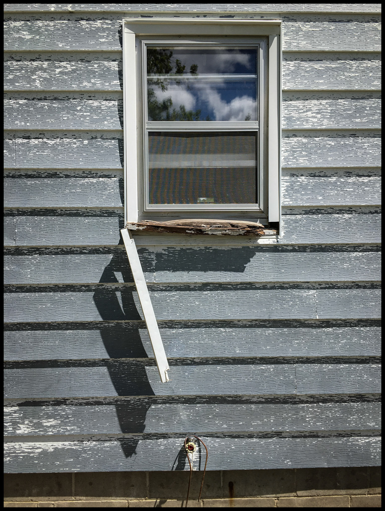 An abstract minimal composition of a piece of fallen trim dangling from a window with a rotted sill on a delapidated house. Kirksville, Missouri 2020