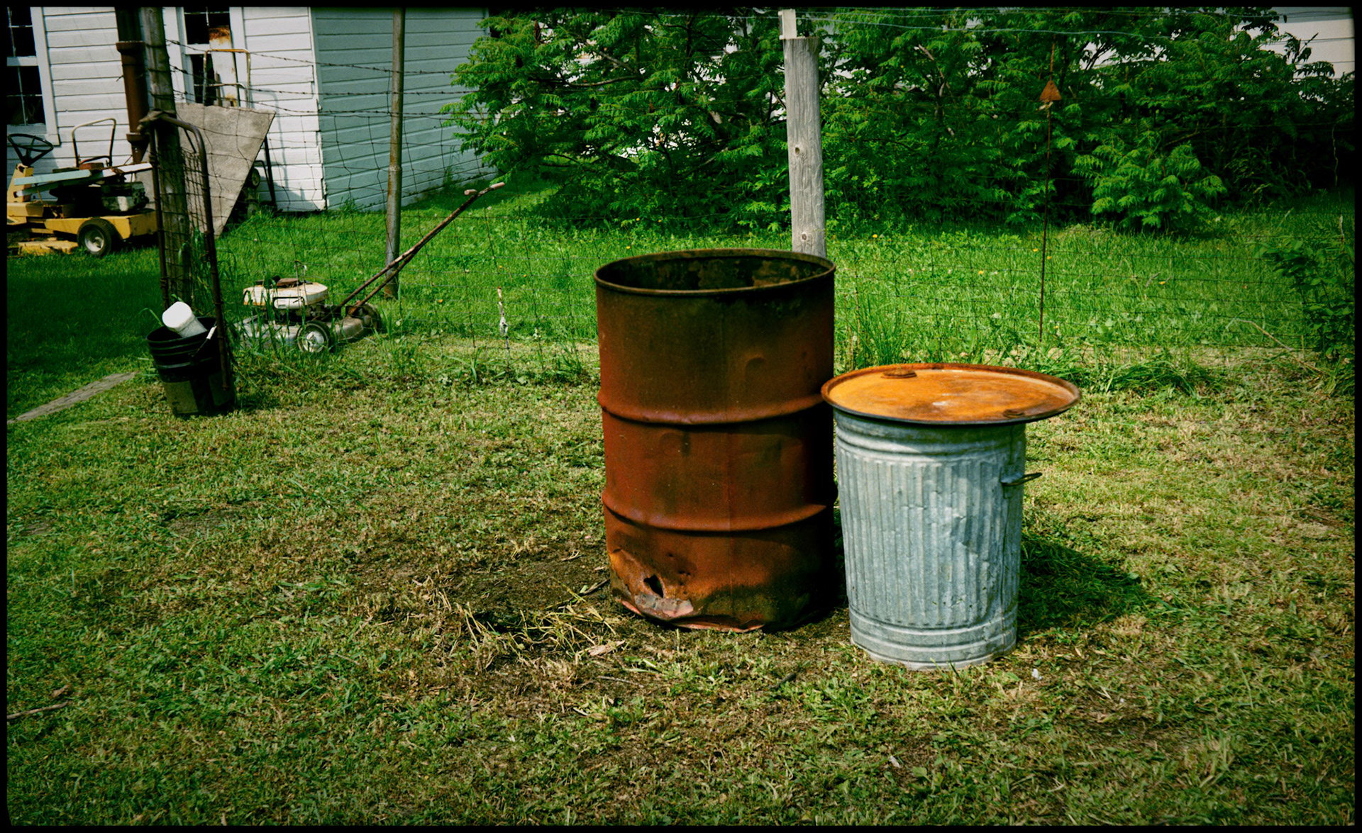 An outdoor still life of a  rusty trash barrel next to a trash can in front of a fence with lawnmowers in the background. Green City, Missouri, USA, 1990.