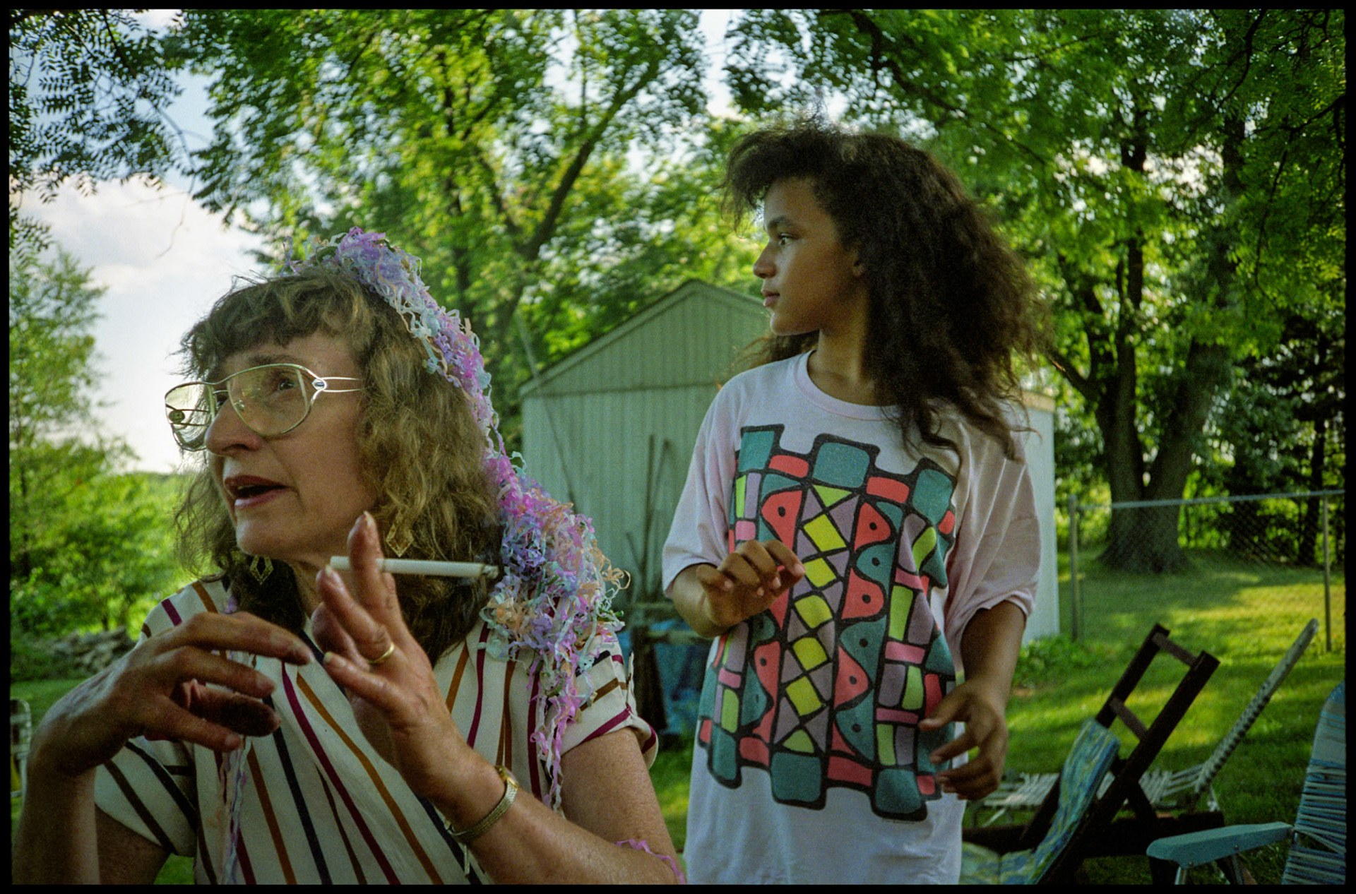 Older woman with cigarette in her hand wearing a headdress made from popper firework streamers on the 4th of July in her back yard as her teenage granddaughter looks on. Near Kansas City, Missouri 1991