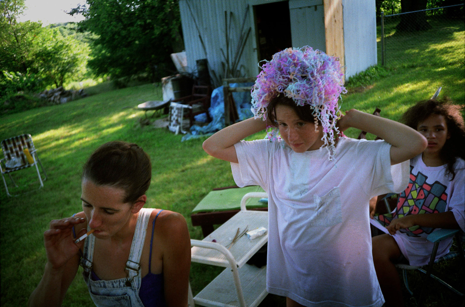 Adolescent girl putting on a headress of made of streamers from popper fireworks on the 4th of July while her mother inhales from a cigarette and her cousin looks on, Near Kansas City, Missouri 1991
