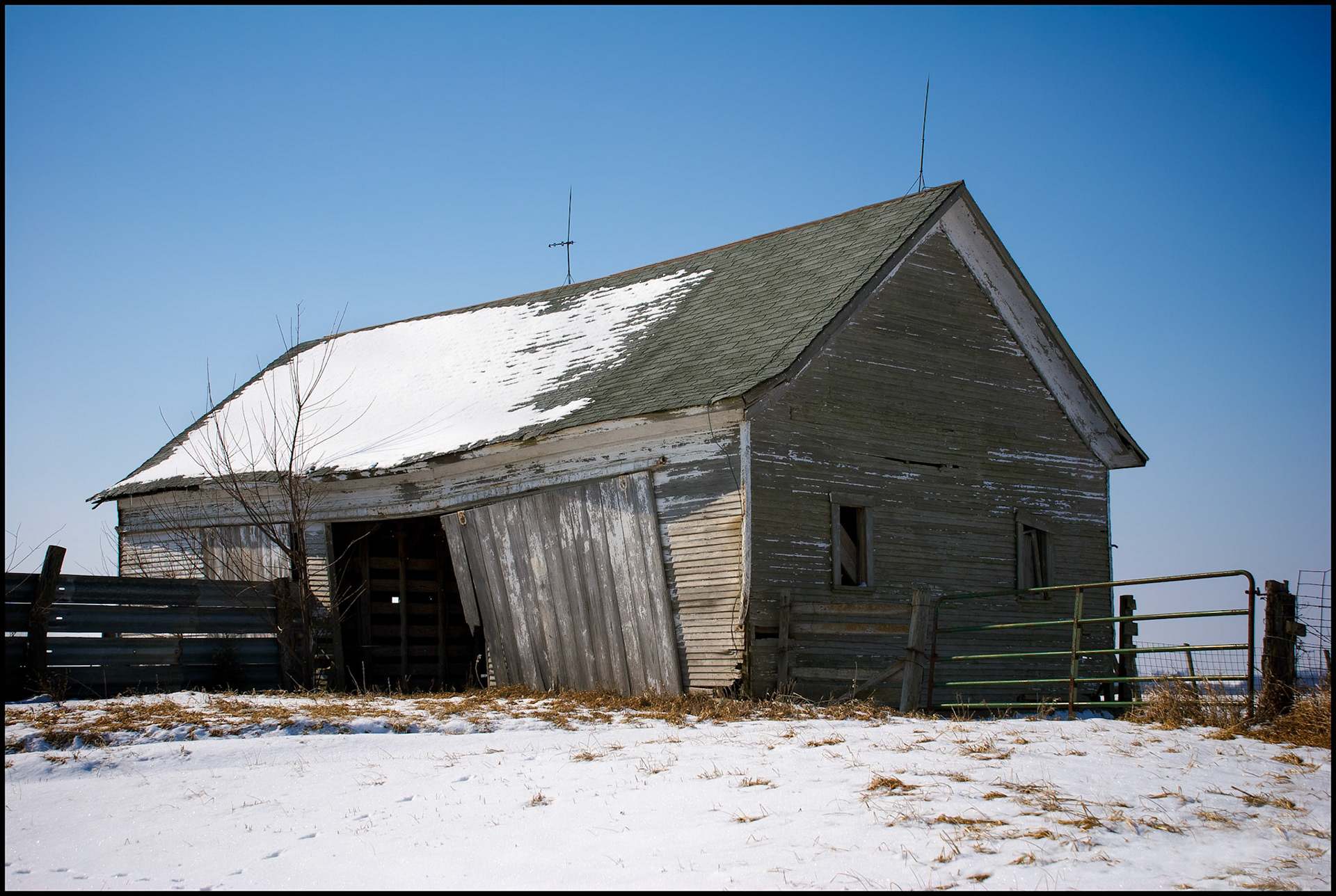 A rural Missouri rustic winter scene of a dilapidated, abandoned barn covered by and surrounded by snow, a fence and gate with a deep blue sky in the background. Near Loeffler, Missouri USA, 2008