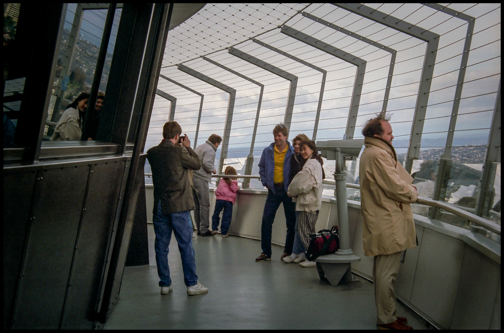 A vintage image of a young man taking a photo of another young man and two young women on the observation deck of the space needle in Seattle, Washington with another older man looking off into the distance in the foreground and another man and his daugther taking in the scene in the background. 1987