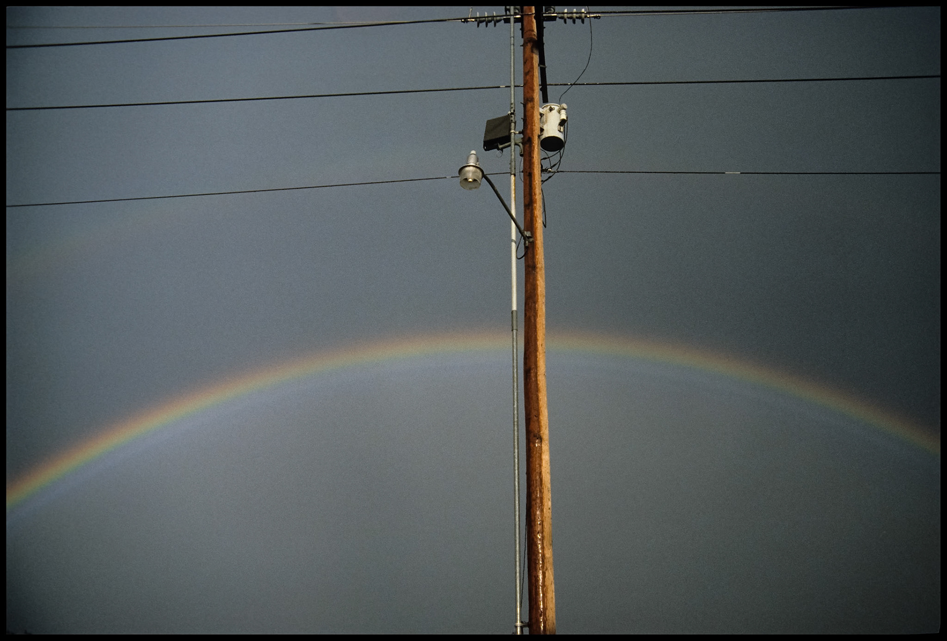 Rainbow behind highlighted power pole with streetlight against a dark grey sky. Kirksville, Missouri 1986