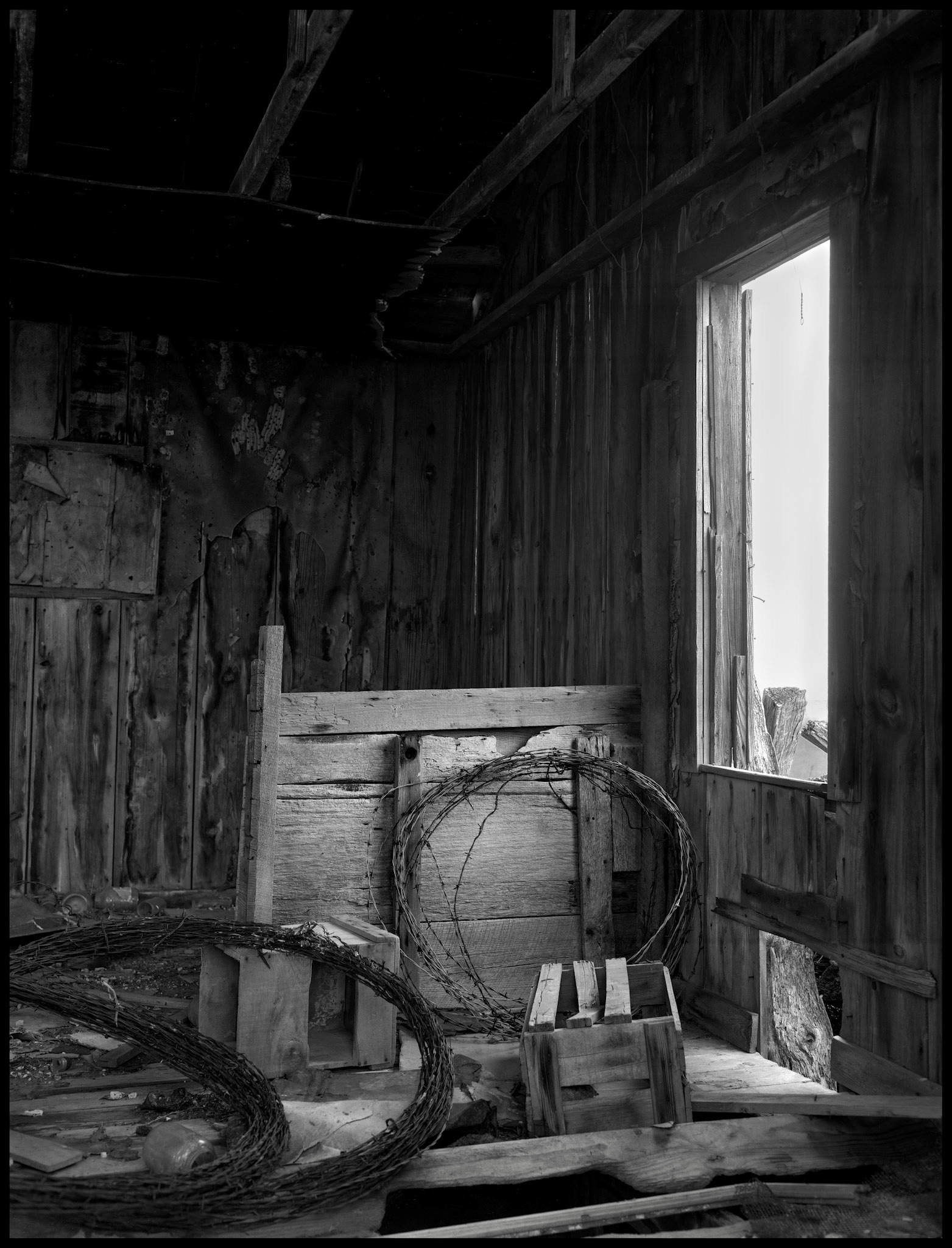 A rustic black and white still life of the interior of an abandoned farm building with spools of barbed wire propped against the former stall wall. Near New Boston, Missouri, 1980