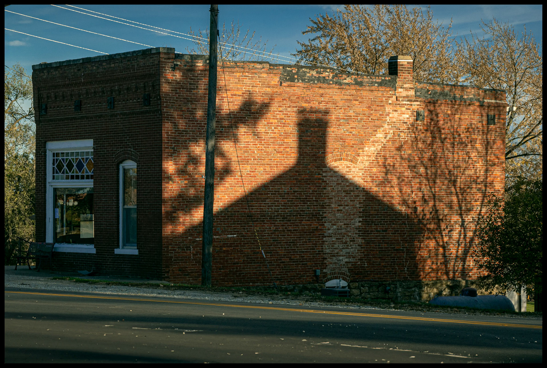 The late Autumn afternoon shadow of a neighboring house and trees blocking the warm afternoon light on the wall of a small town post office. Ethel, Missouri 2023