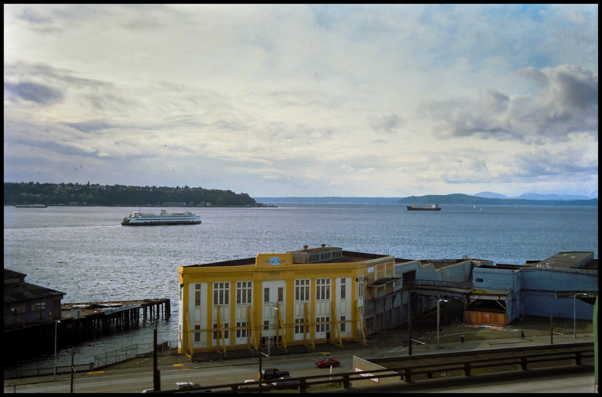 An historic view of Puget Sound with the historic Pier 64 Port of Seattle building in the foreground and a ferry and barge on the water at the Seattle waterfront. Seattle, Washington 1987