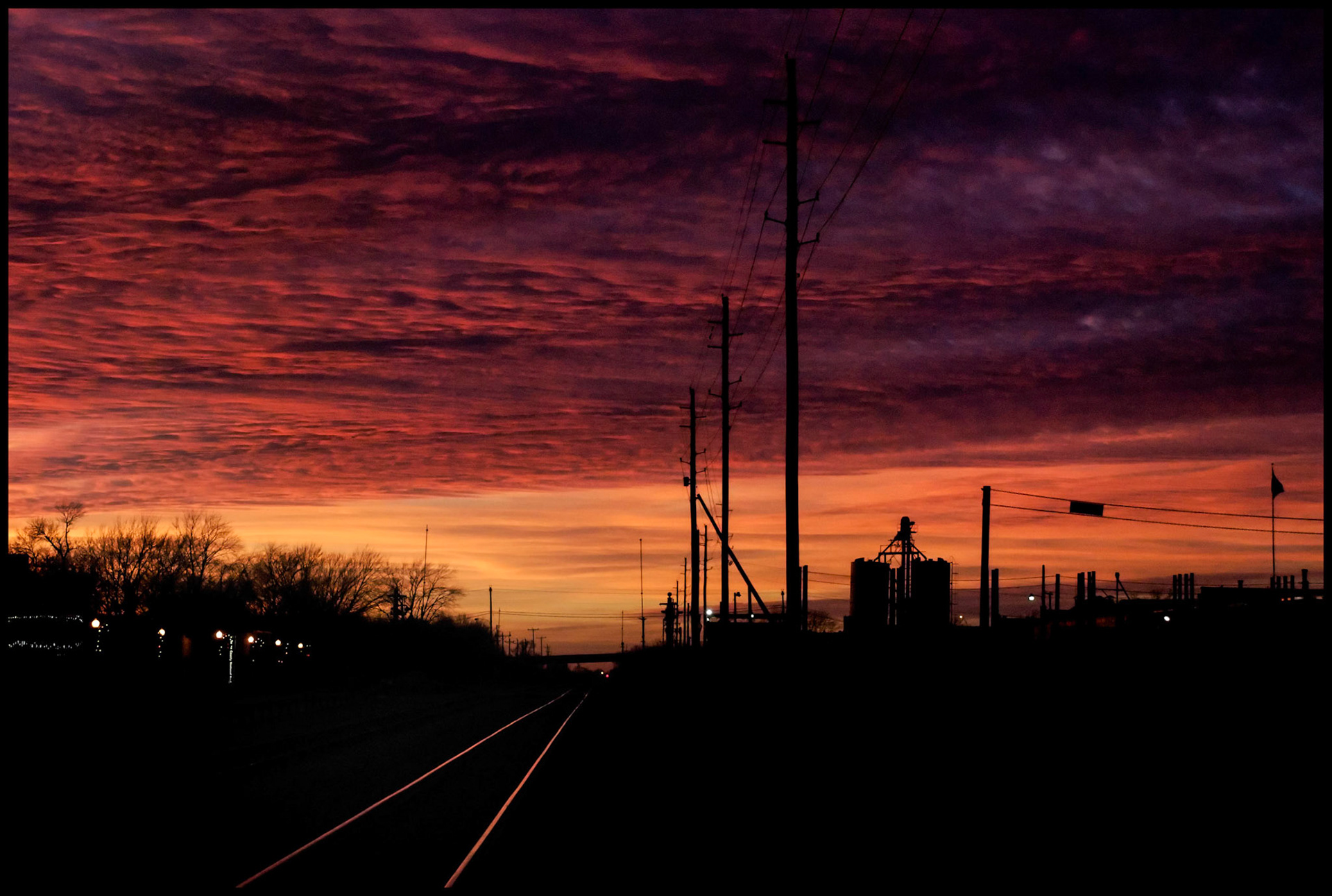 Textured Orange and Pink clouds at sunset behind silhouetted grain elevator and power poles in a small midwest town with highlight on railroad tracks in foreground leading off into the distance. Centralia, Missouri USA 2008